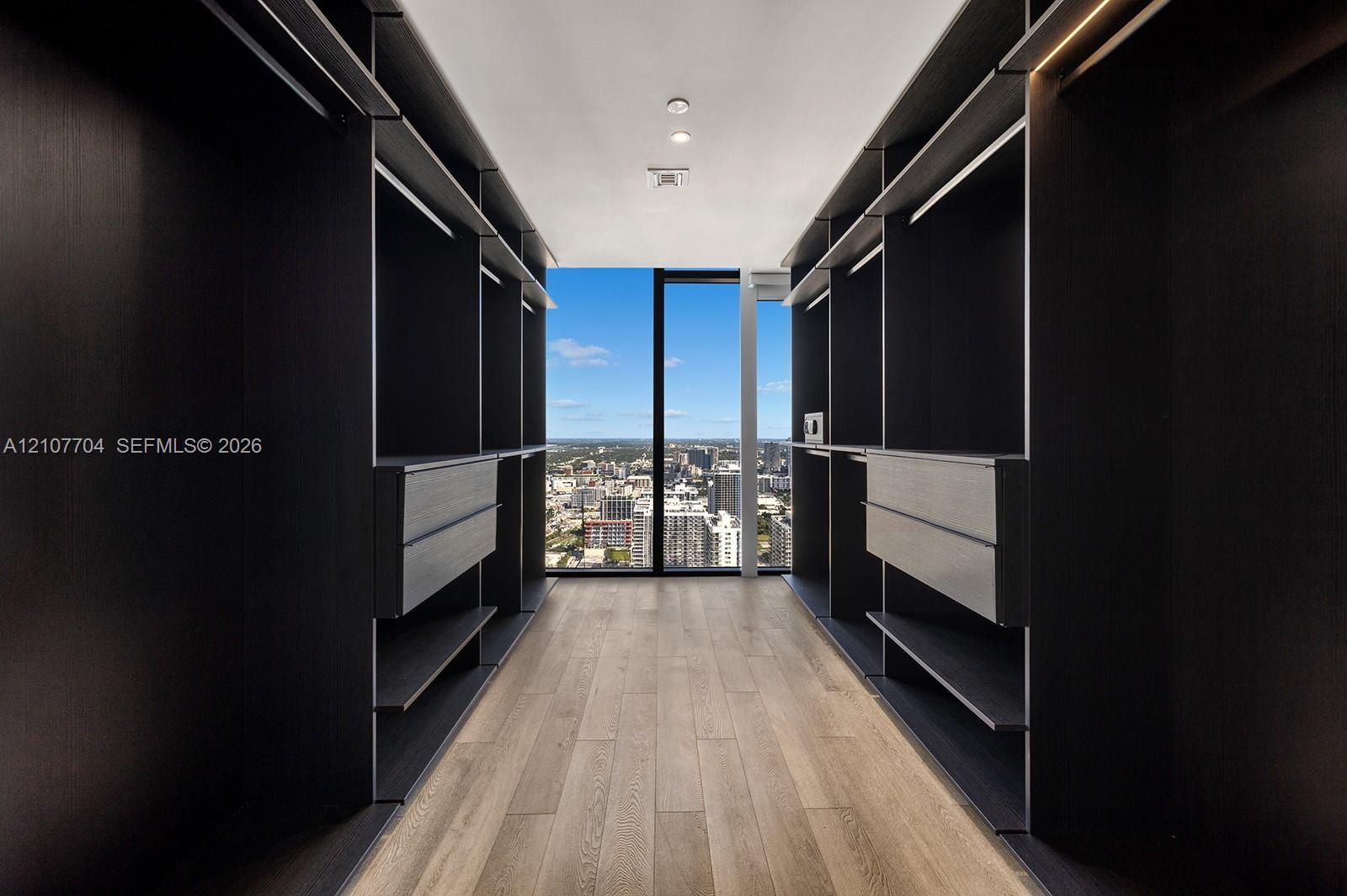 Custom walk-in closet featuring floor-to-ceiling dark cabinetry, integrated shelving, drawer units, wood-finish flooring, and full-height window panel