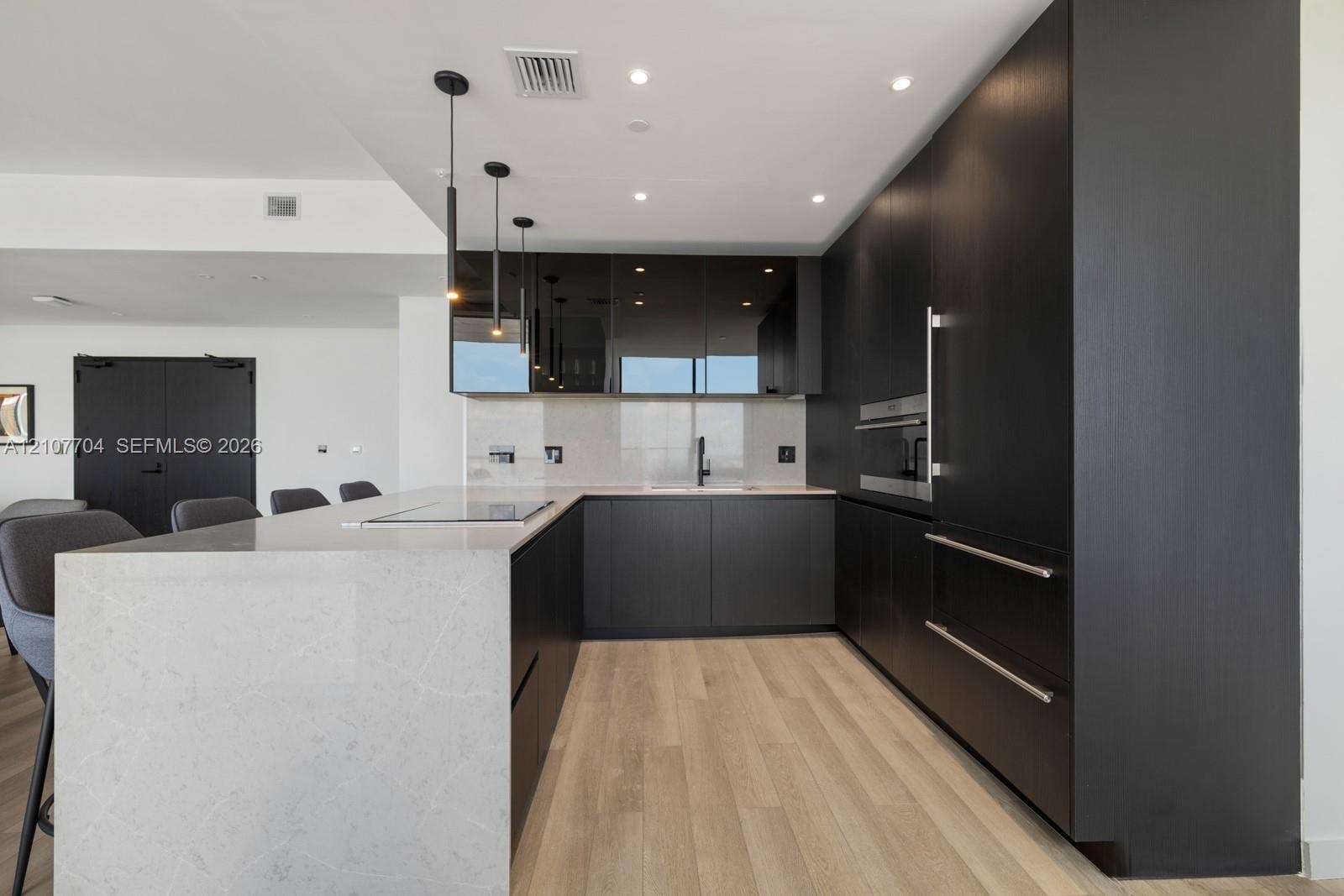 Contemporary kitchen featuring custom dark cabinetry, stone-surfaced waterfall island, integrated built-in appliances, recessed lighting, and wood-fin