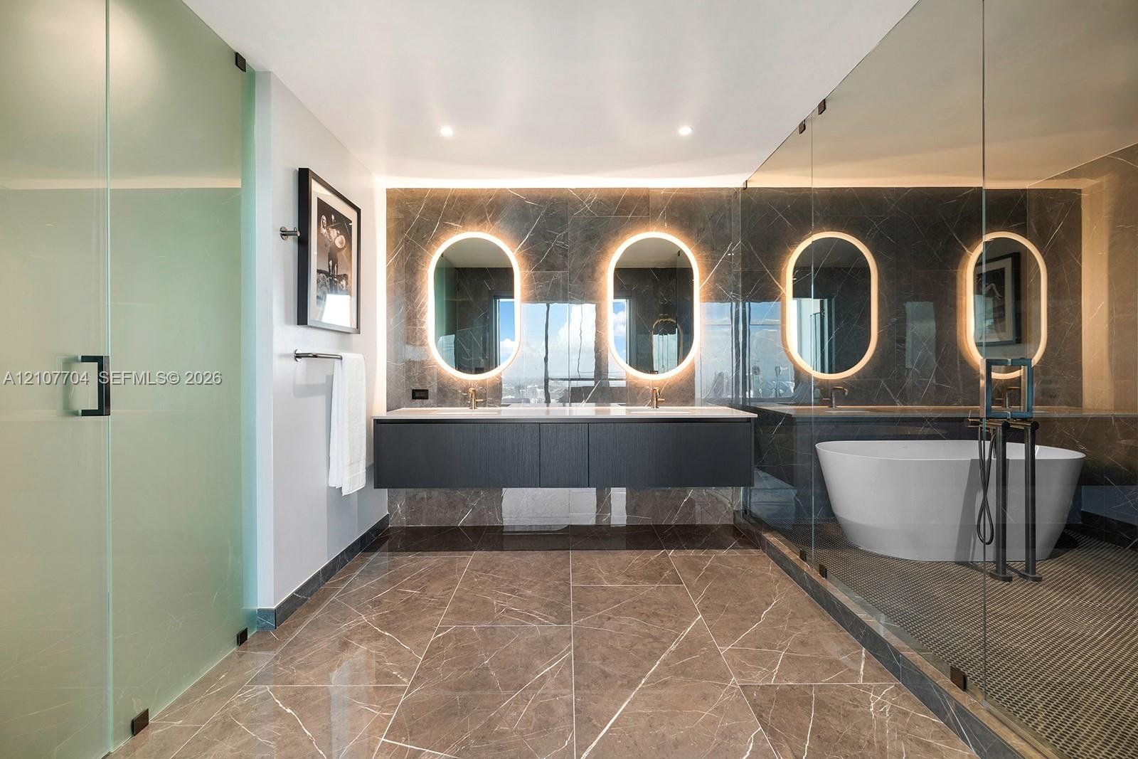 Floating dark wood-finish vanity with dual sinks, backlit oval mirrors, stone-tiled walls and flooring, glass-enclosed walk-in shower, and freestandin