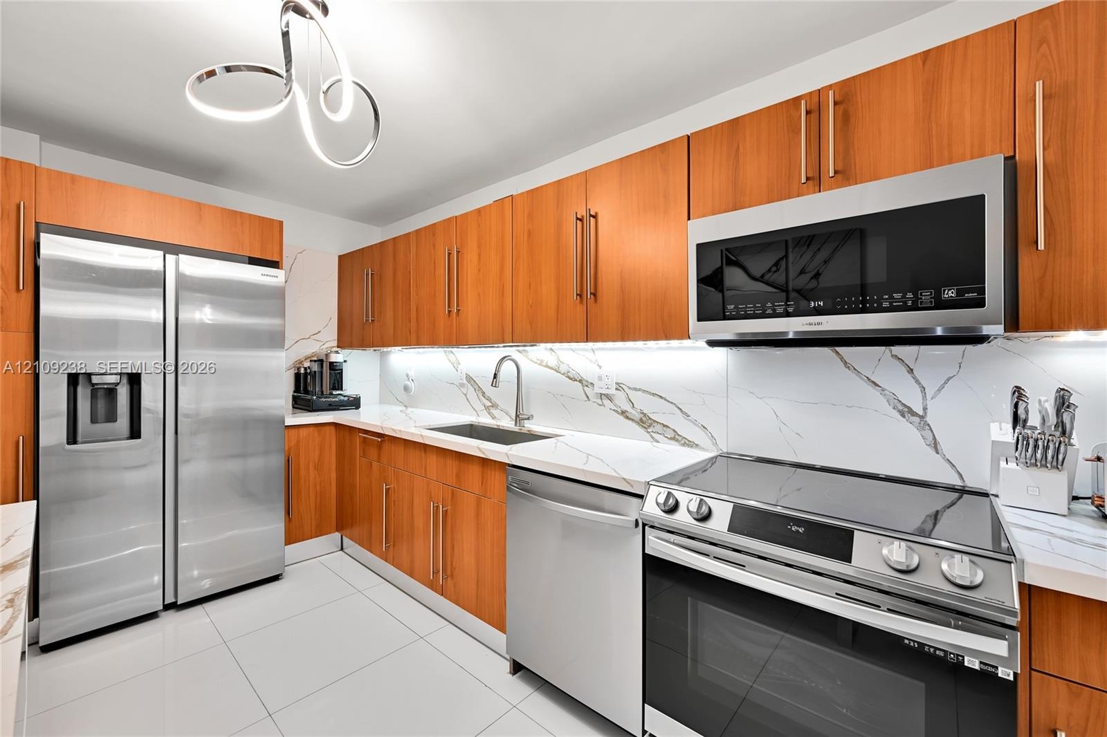 Kitchen featuring wood-finish cabinetry, stone-patterned backsplash, stainless steel appliances, under-cabinet lighting, and large-format tile floorin