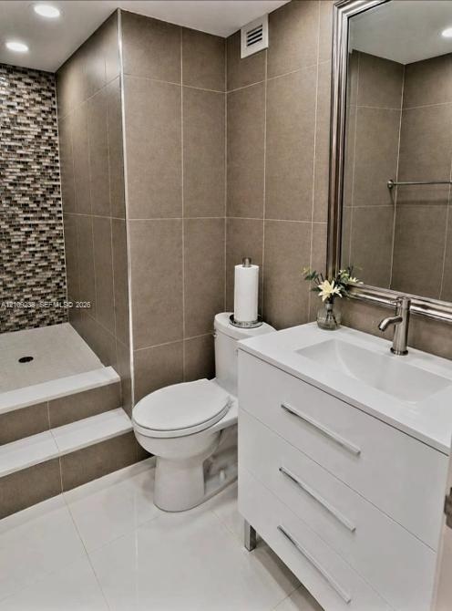 Bathroom featuring a modern white vanity, glass-tiled shower wall accent, large-format neutral wall tiling, stepped shower entry, and a framed mirror