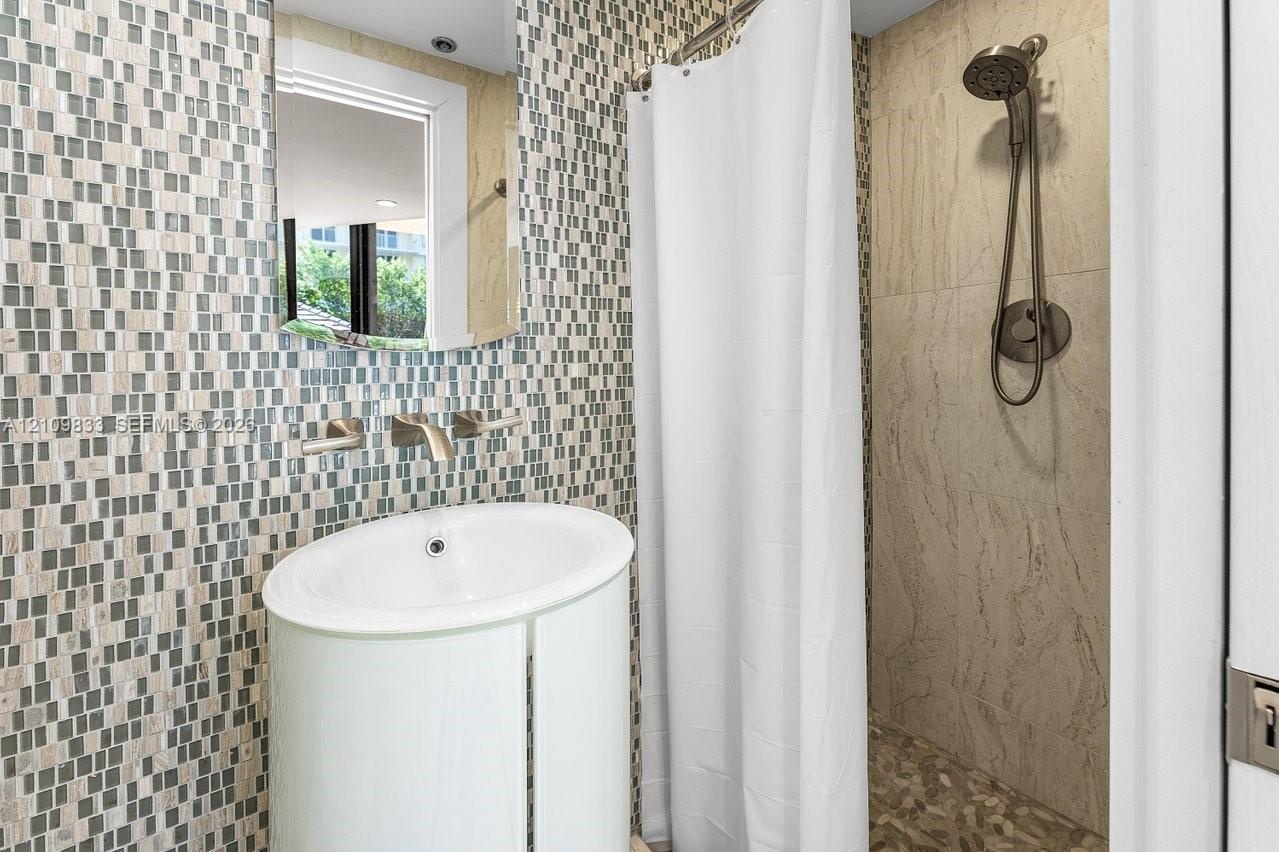 Bathroom featuring a pedestal sink, wall-mounted faucet, mosaic tile backsplash, walk-in shower with stone tile walls, and handheld shower fixture