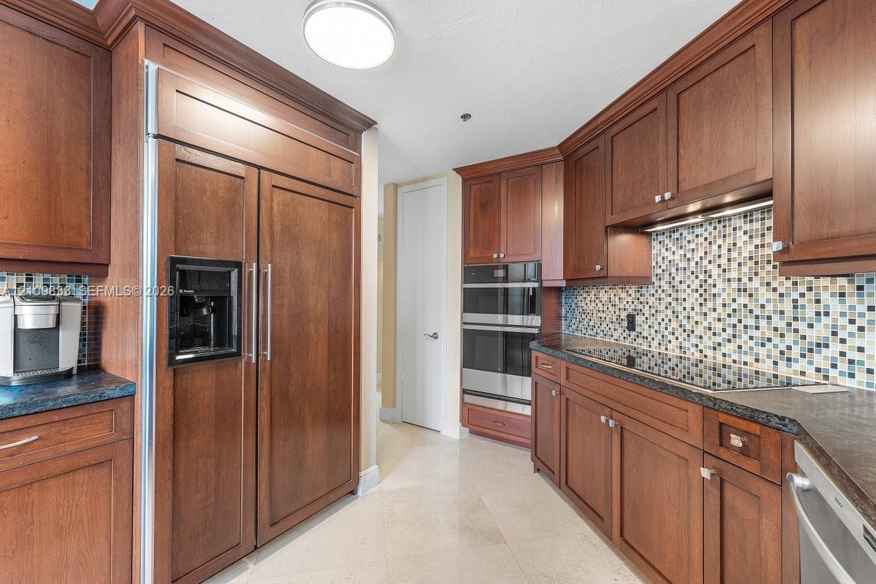 Kitchen featuring dark wood cabinetry, panel-front refrigerator, stainless steel built-in double ovens, multi-colored glass tile backsplash, and stone