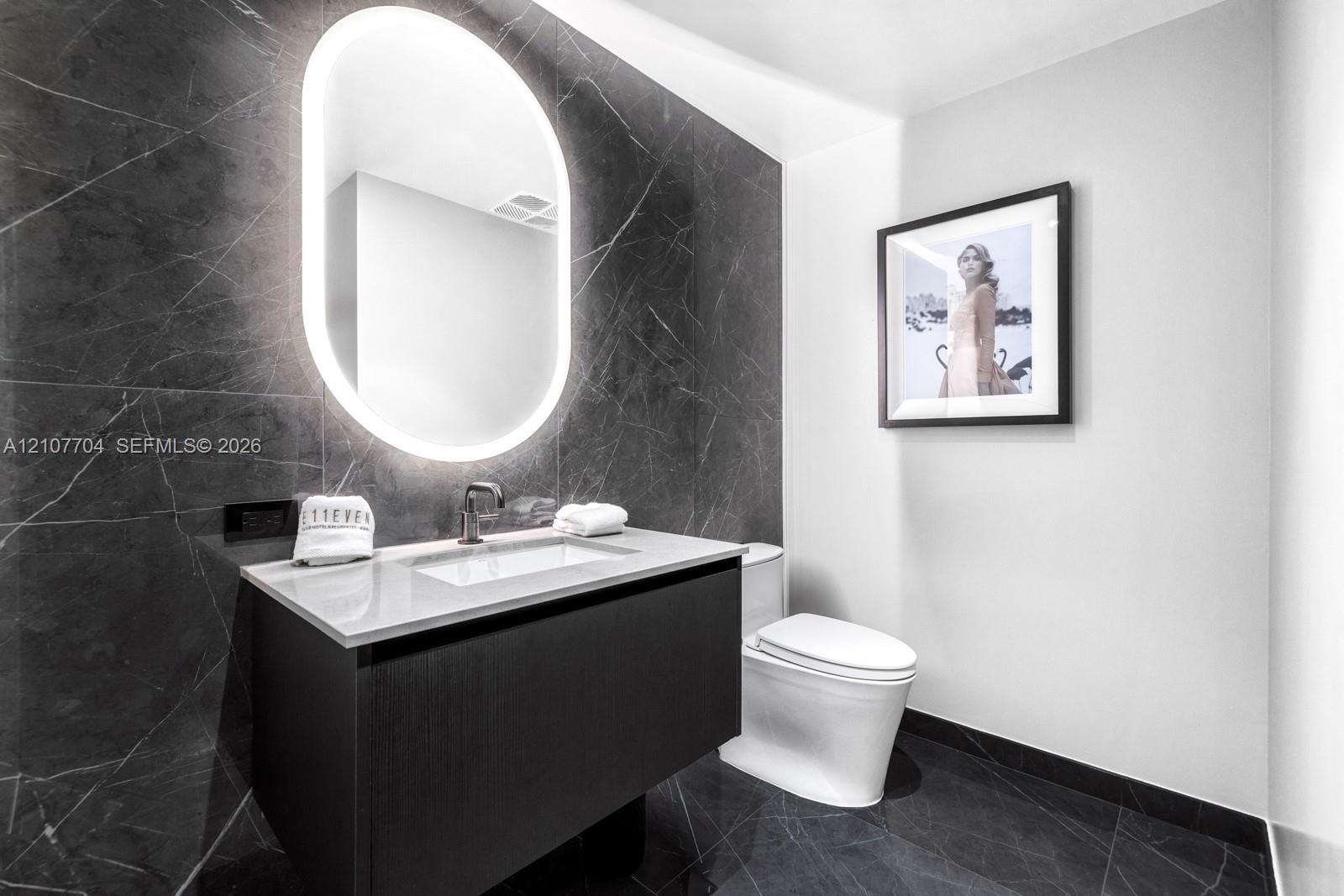 Floating vanity with a light-toned countertop, dark stone-finish wall tile, integrated oval back-lit mirror, and modern wall-mounted faucet