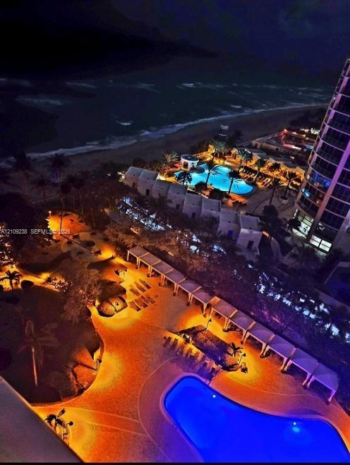 Aerial perspective of oceanfront property featuring a multi-level swimming pool, expansive paved terrace, linear cabanas, and manicured grounds