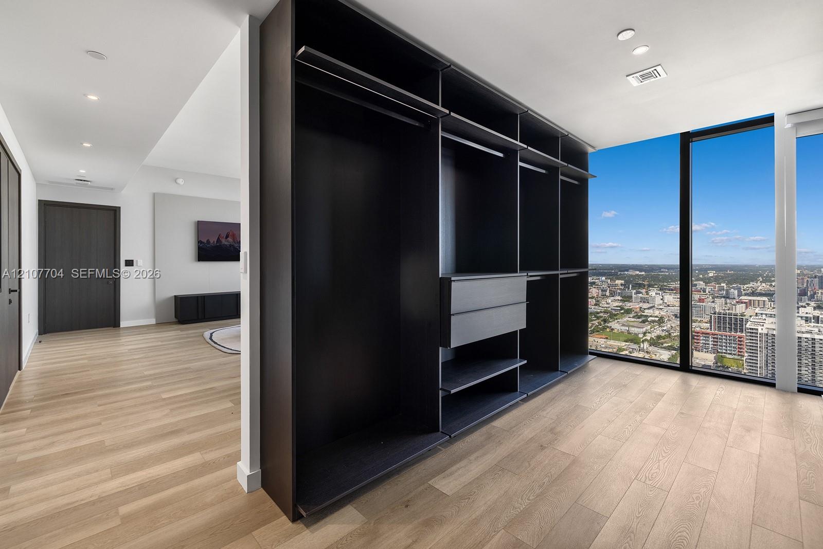 Custom-built dark finish storage cabinetry featuring integrated drawers and hanging rods, light-toned wood-finish flooring, and floor-to-ceiling windo