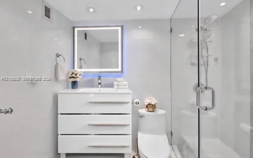 Bathroom featuring a white three-drawer vanity, illuminated rectangular mirror, one-piece elongated toilet, and a glass-enclosed shower with polished 