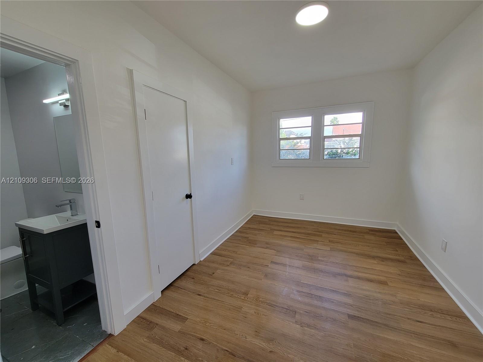 Interior space featuring wood-finish flooring, white walls, dual-pane windows, and a recessed ceiling light, accompanied by a bathroom with a charcoal