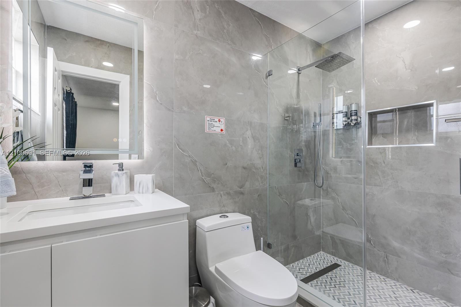 Bathroom featuring large-format gray tile wall surrounds, a frameless glass shower enclosure with a rainfall showerhead, a built-in tiled shower niche
