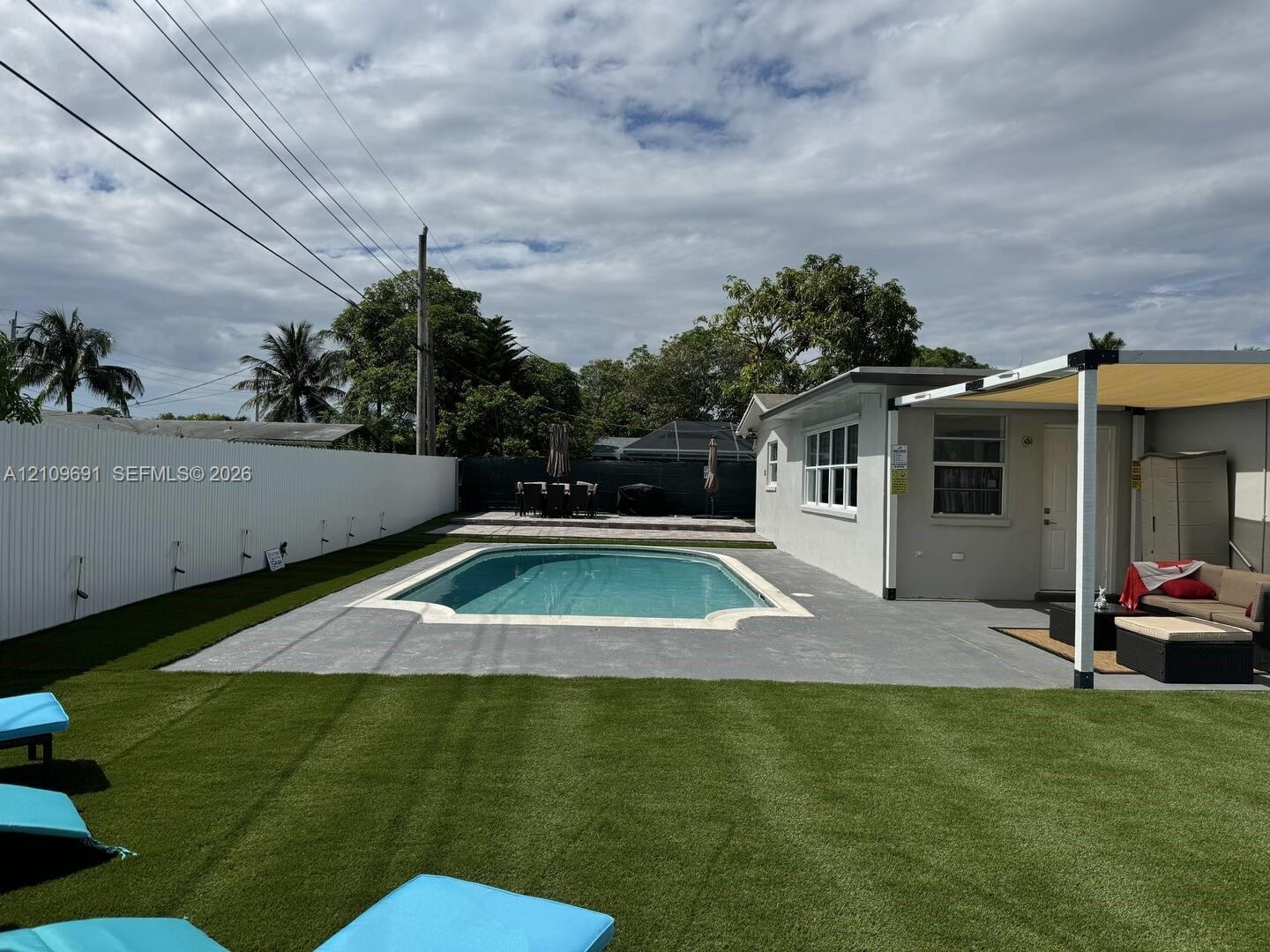 In-ground pool with concrete surround, synthetic turf lawn, white privacy fencing, and attached patio cover