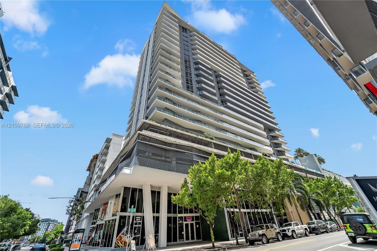 High-rise building featuring a glass facade, multiple cantilevered balconies, decorative geometric screening, and ground-level storefronts with floor-
