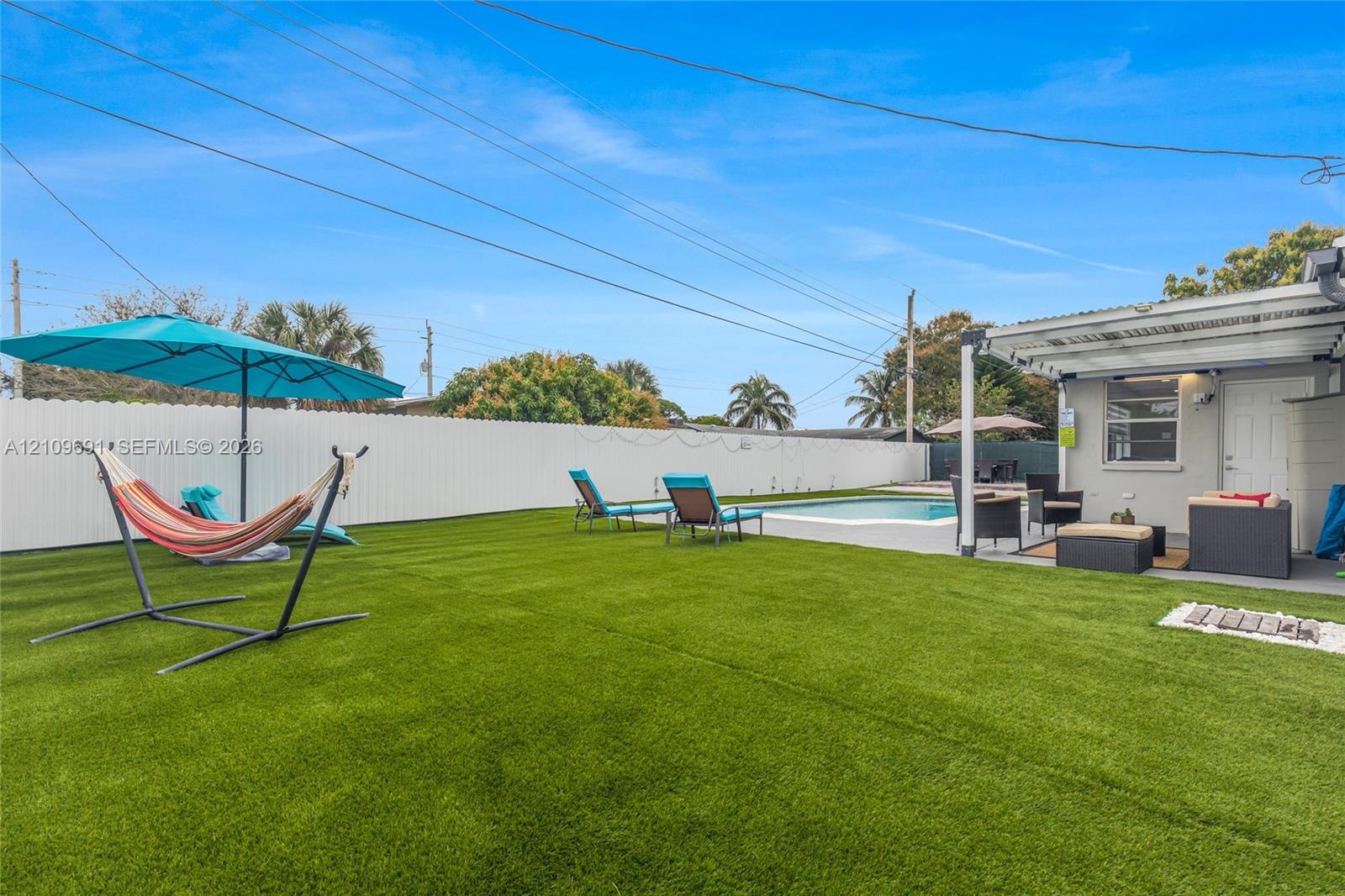 Backyard featuring a synthetic turf lawn, in-ground swimming pool with white masonry surround, white vinyl privacy fencing, and a metal-covered patio 