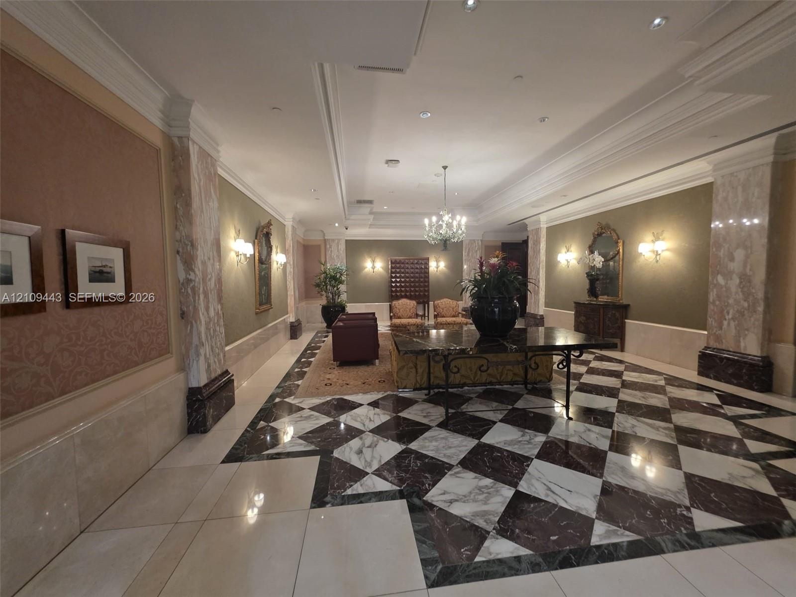 Grand entrance hall featuring patterned stone flooring, decorative stone-clad columns, recessed ceiling lighting, wall-mounted sconces, and crown mold