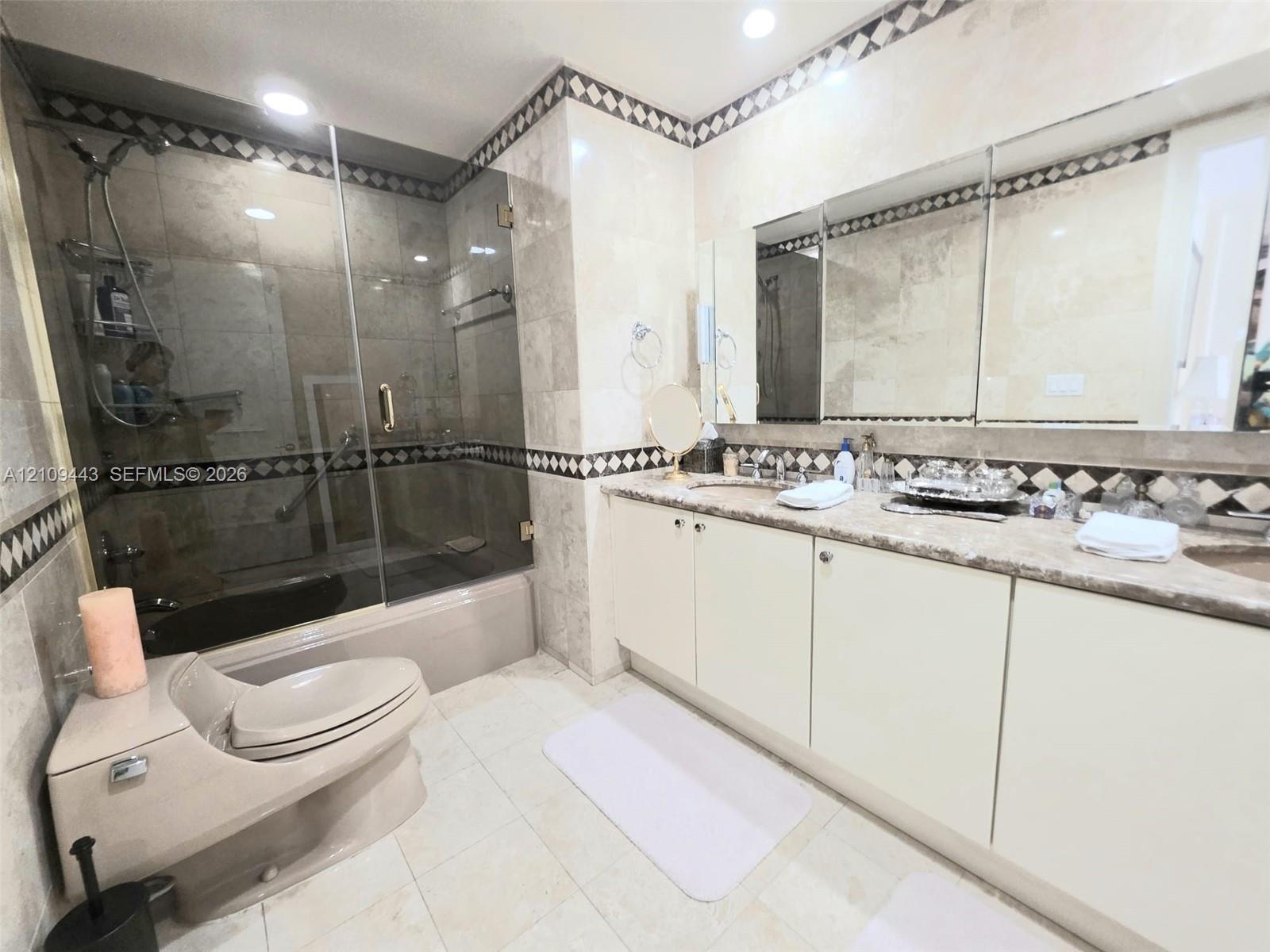Bathroom featuring stone-tiled walls with decorative mosaic trim, dual-sink vanity with solid-surface countertop, frameless glass shower enclosure, an