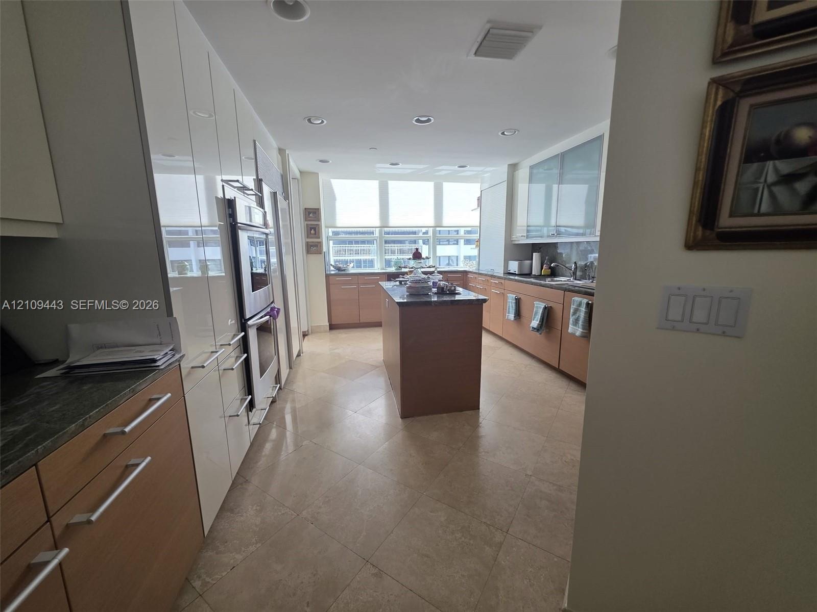 Kitchen featuring two-tone cabinetry with wood-finish lower units and high-gloss upper cabinetry, stone countertops, a central kitchen island, and lar