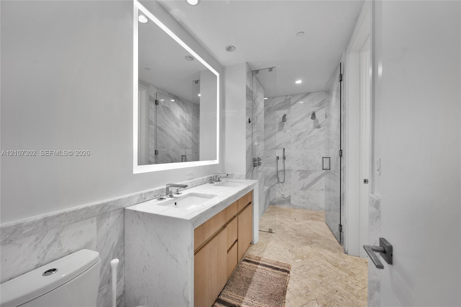 Dual-vanity cabinetry with stone countertop, integrated light-up mirror, walk-in shower with stone wall tile, and herringbone-patterned stone flooring