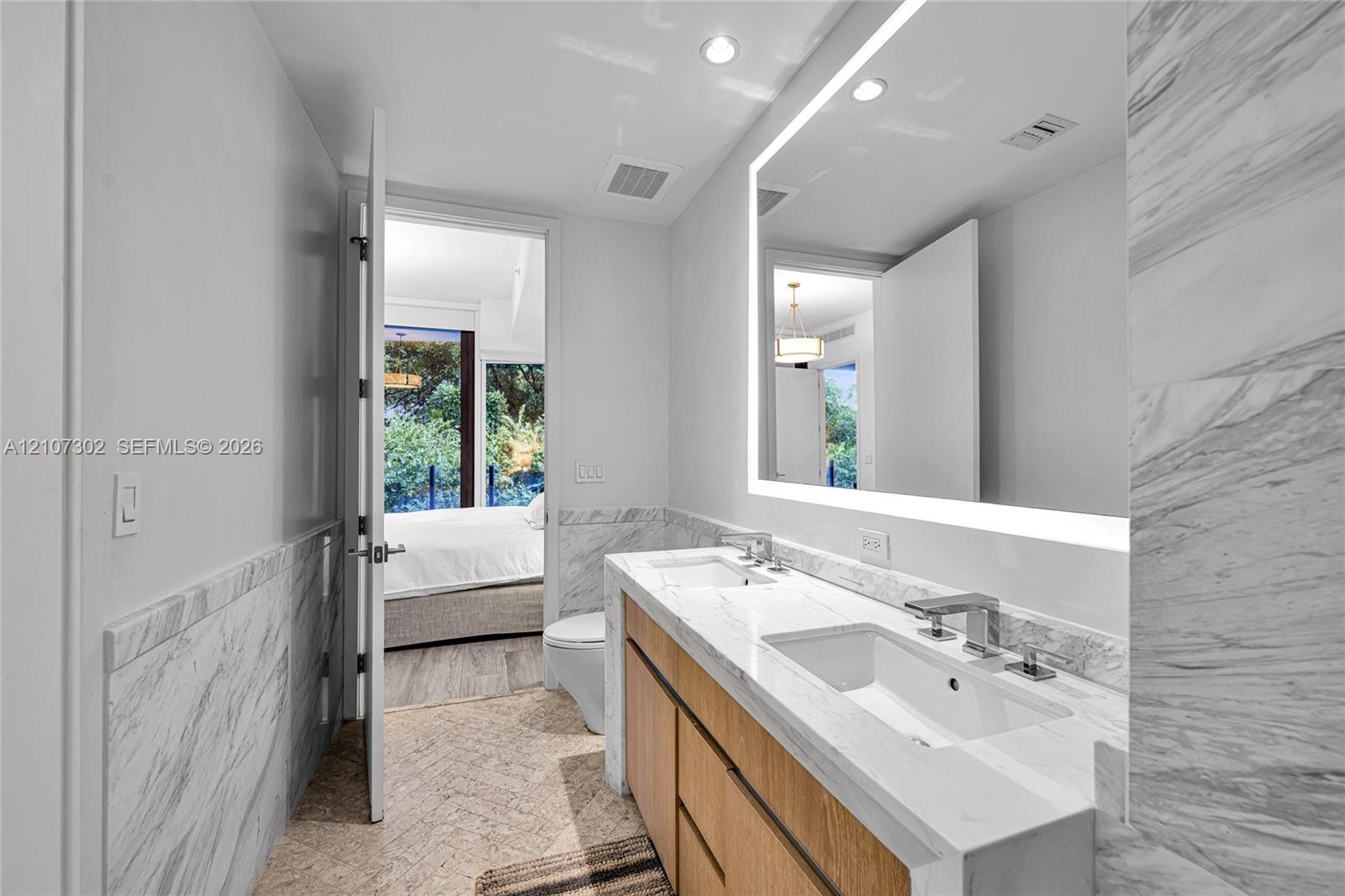 Bathroom featuring a dual-sink vanity with stone countertops, a large LED-backlit mirror, and stone wall tiling
