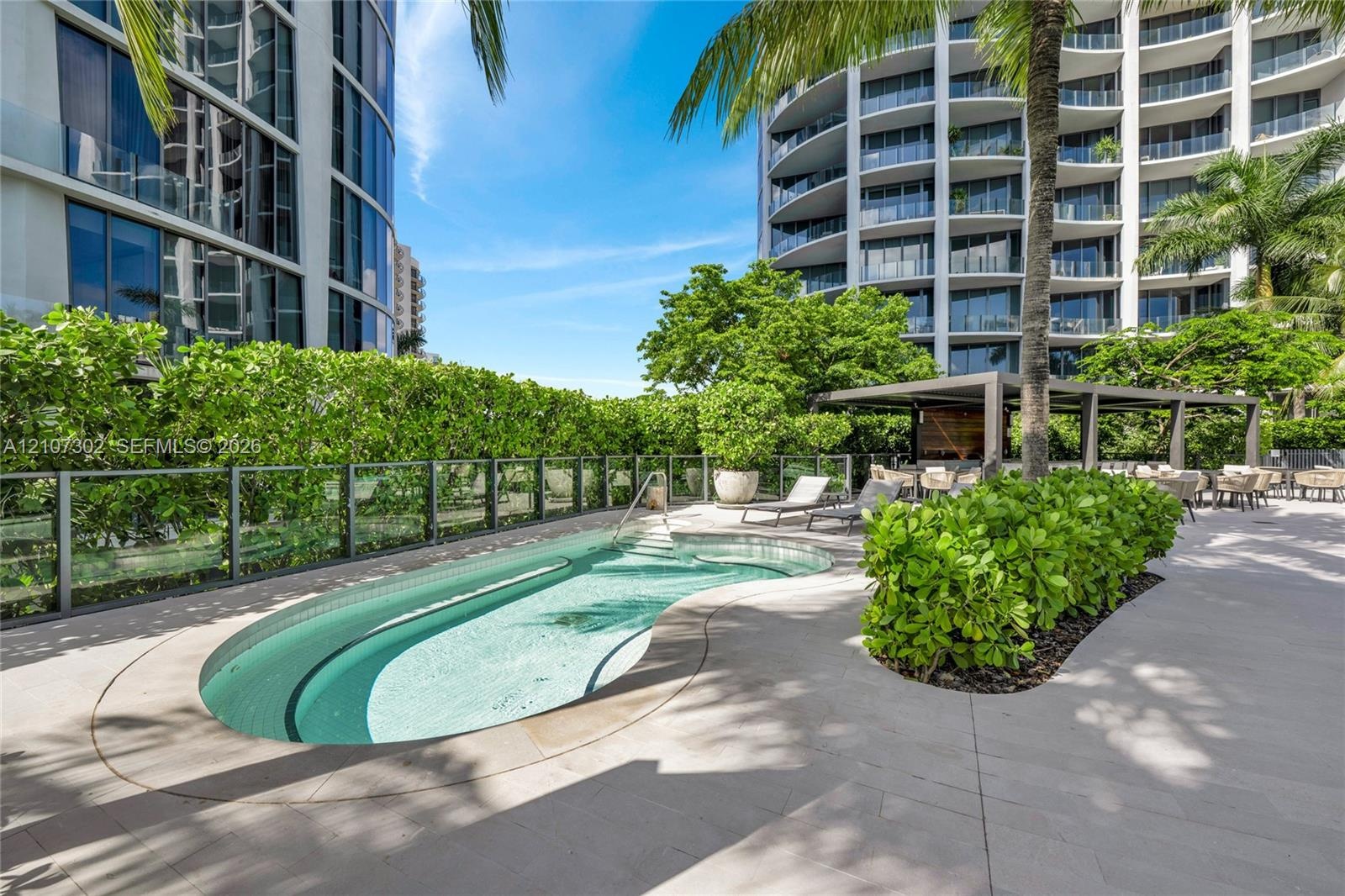 Curved swimming pool featuring a glass pane perimeter fence, stone-finish decking, and a covered pavilion structure