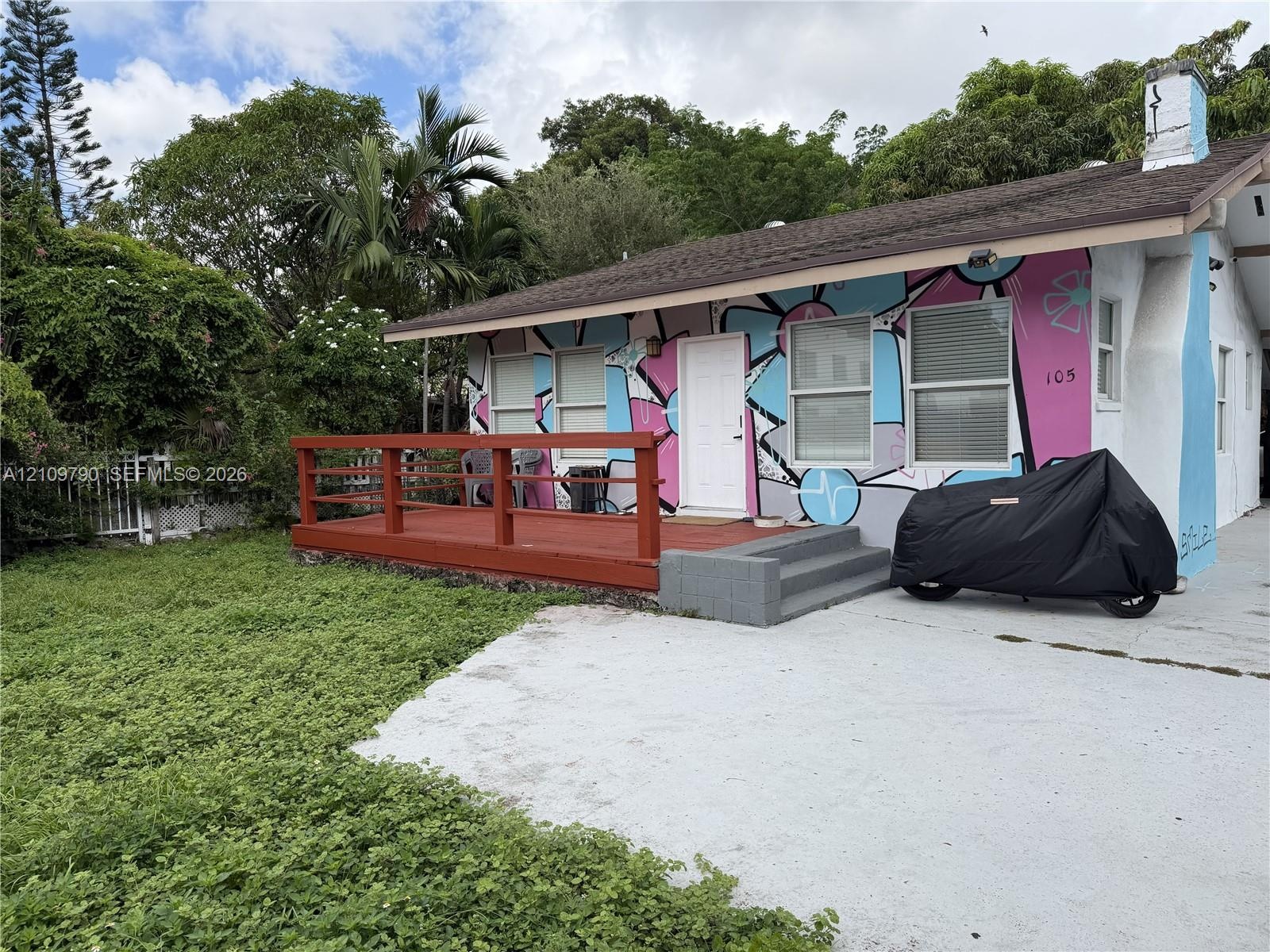Single-story residence featuring an exterior mural, raised wood-finish deck with railing, shingle roof, concrete driveway, and mature landscaping