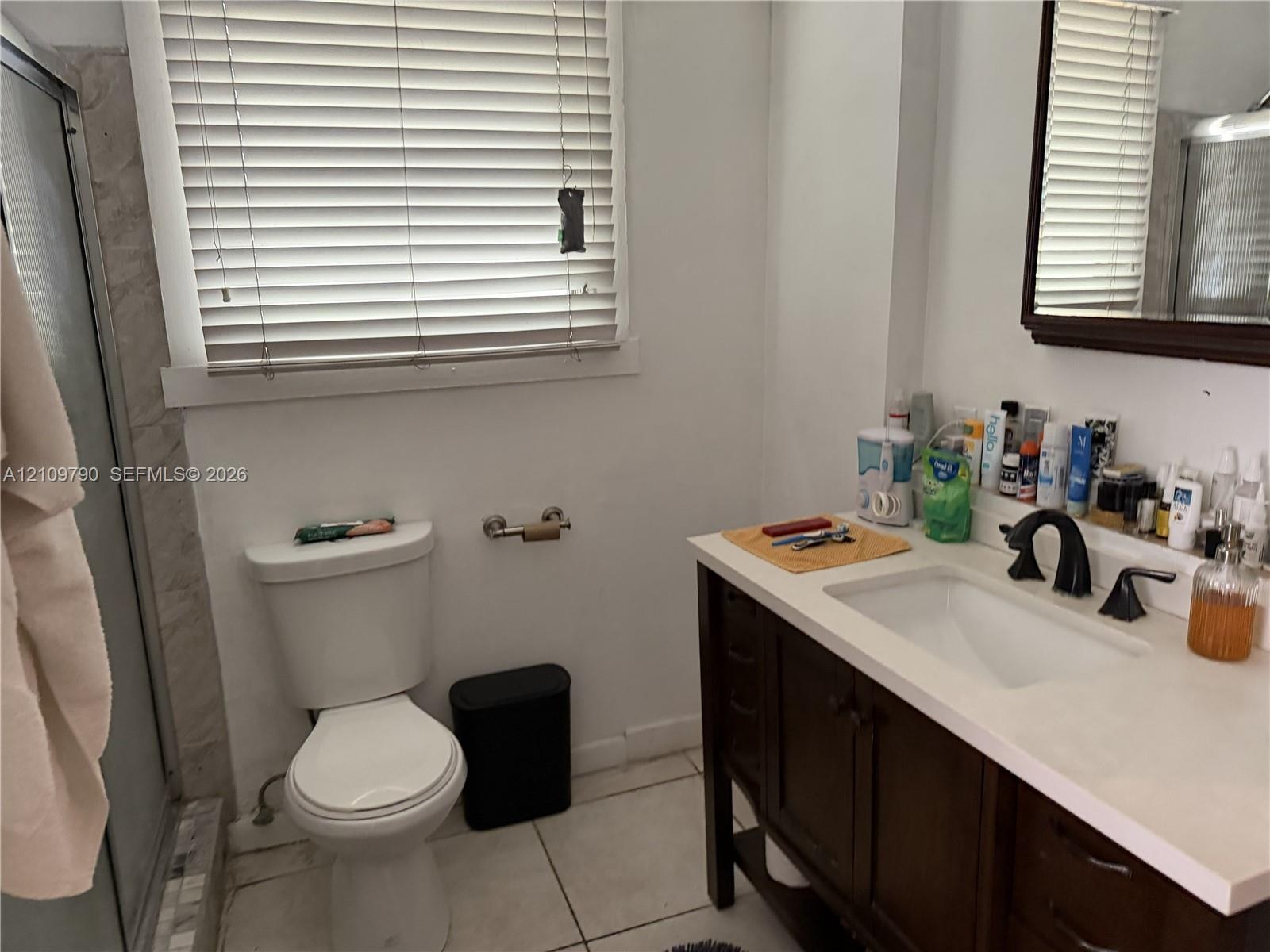 Bathroom featuring a dark wood-finish vanity, light-toned stone countertop, undermount sink with dark metal hardware, ceramic floor tile, and shower e