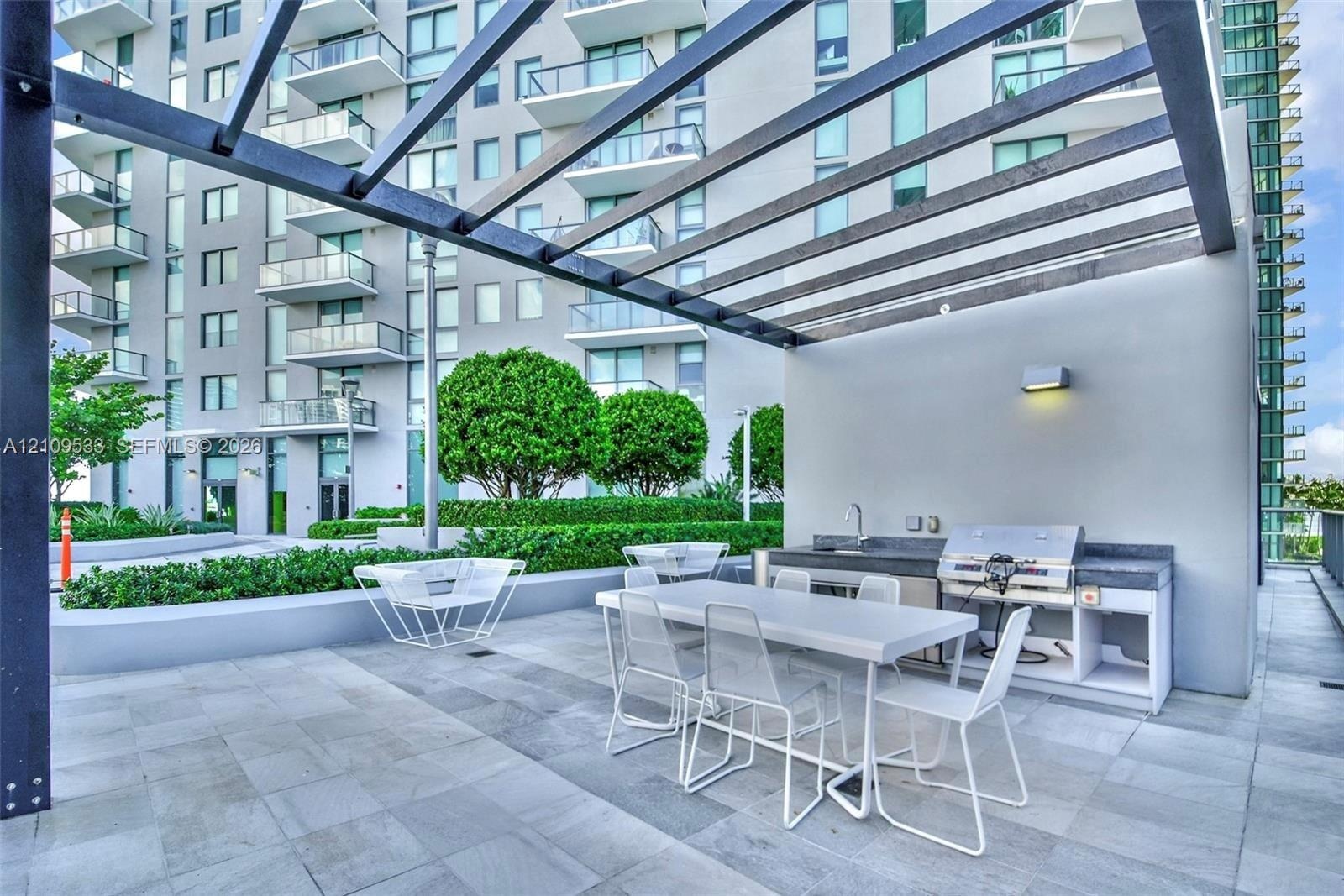 Outdoor terrace featuring an overhead pergola, built-in kitchen station with a stainless steel grill and sink, stone-finish flooring, and manicured la