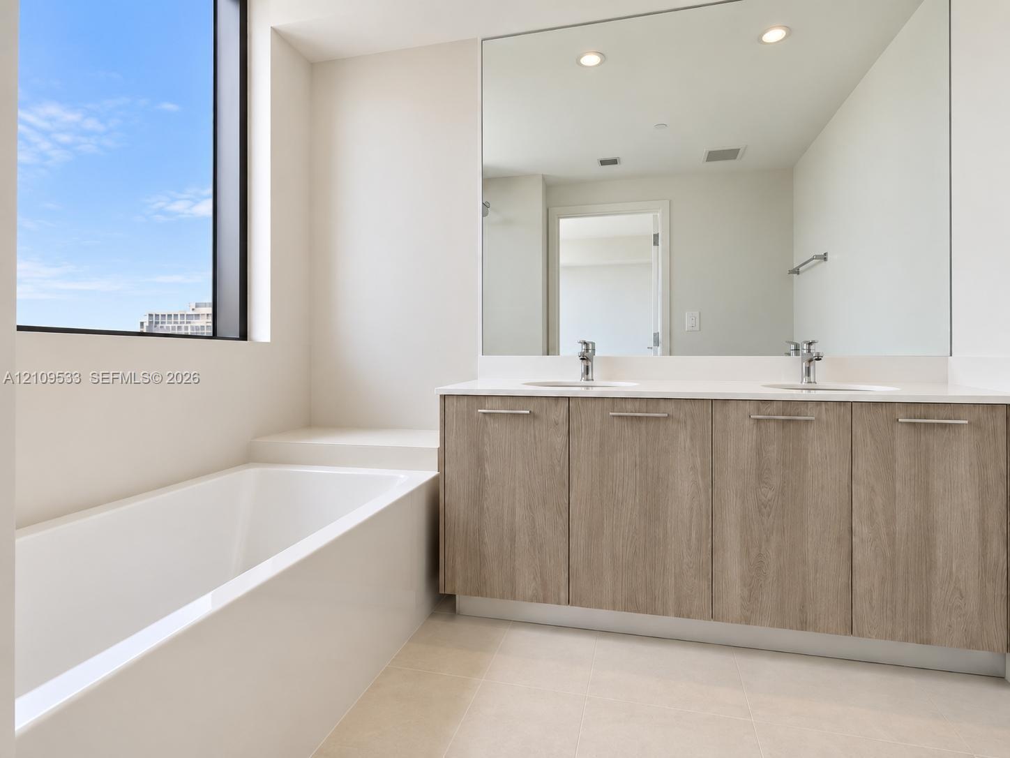 Bathroom featuring a double-sink vanity with wood-finish cabinetry, a white countertop, an integrated soaking tub, and a floor-to-ceiling window