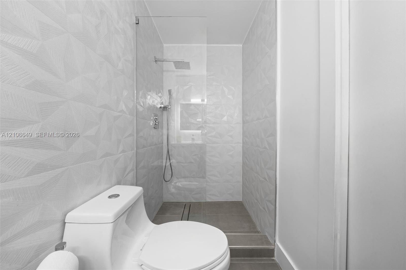 Walk-in shower featuring textured geometric wall tile, a frameless glass enclosure, a rainfall showerhead, and a built-in recessed niche