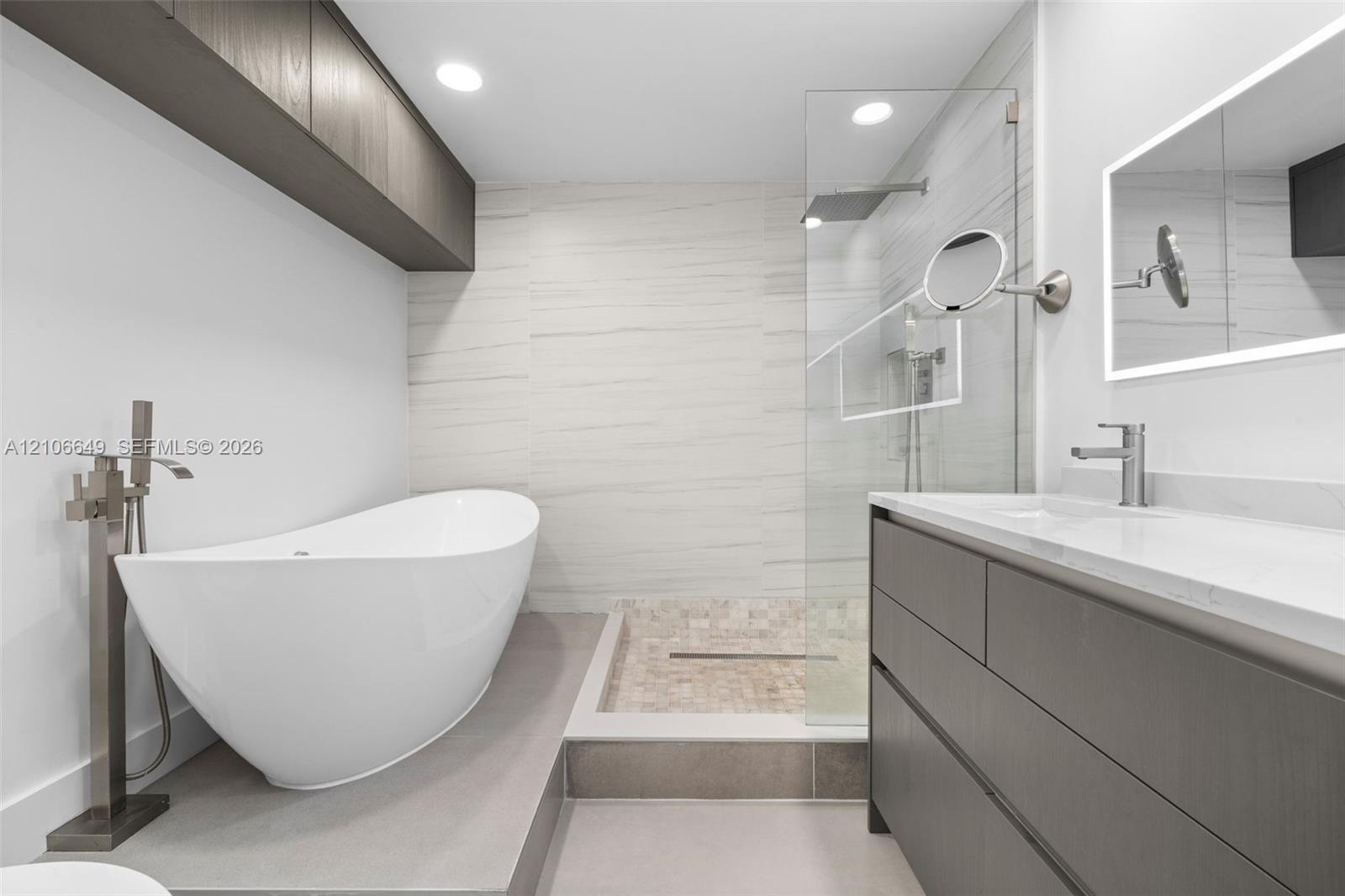 Freestanding soaking tub with floor-mounted faucet, walk-in glass-enclosed shower featuring mosaic stone flooring, dark wood-finish cabinetry, illumin