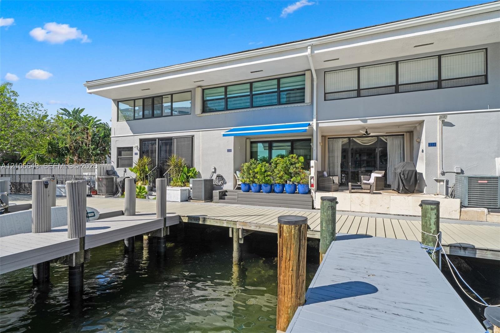Waterfront exterior featuring a multi-level composite boat dock, stone-surfaced patio area, retractable blue awning, and large-format sliding glass do