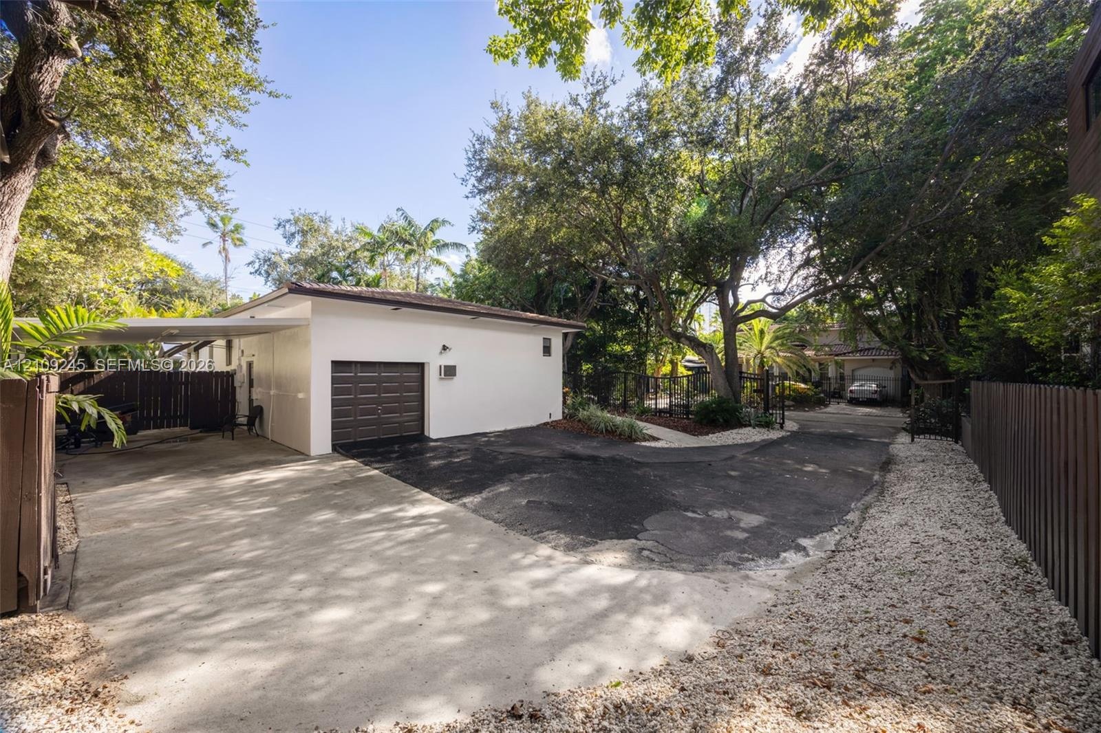 Detached garage with a multi-vehicle driveway, covered carport, mature tree canopy, and wood-slat perimeter fencing