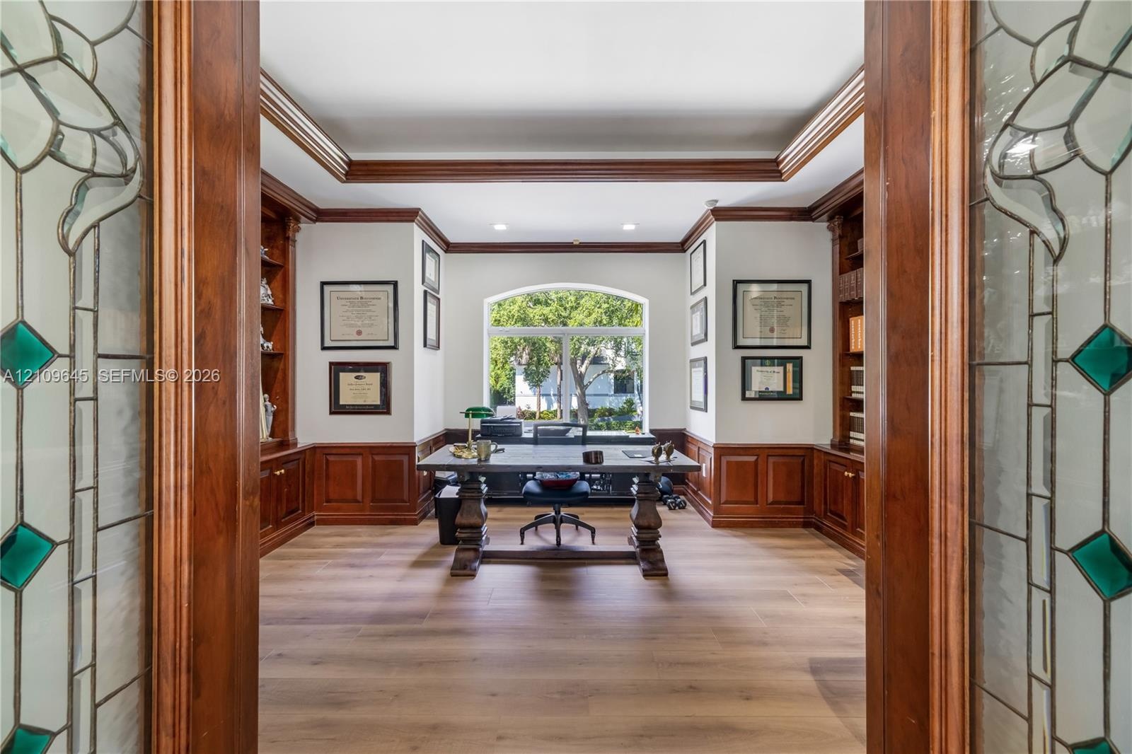Custom woodwork study featuring wood-paneled wainscoting, floor-to-ceiling built-in cabinetry, arched window, recessed lighting, and glass-paneled dou