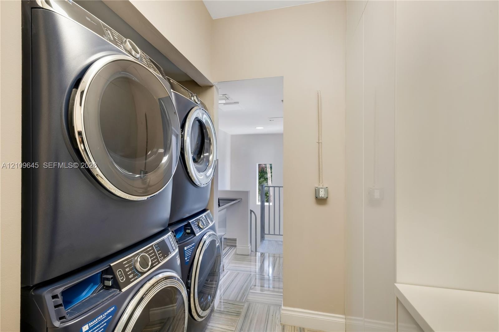 Laundry area featuring stacked metallic-finish washing machine and dryer, floor-to-ceiling built-in cabinetry, and neutral stone-patterned tile floori