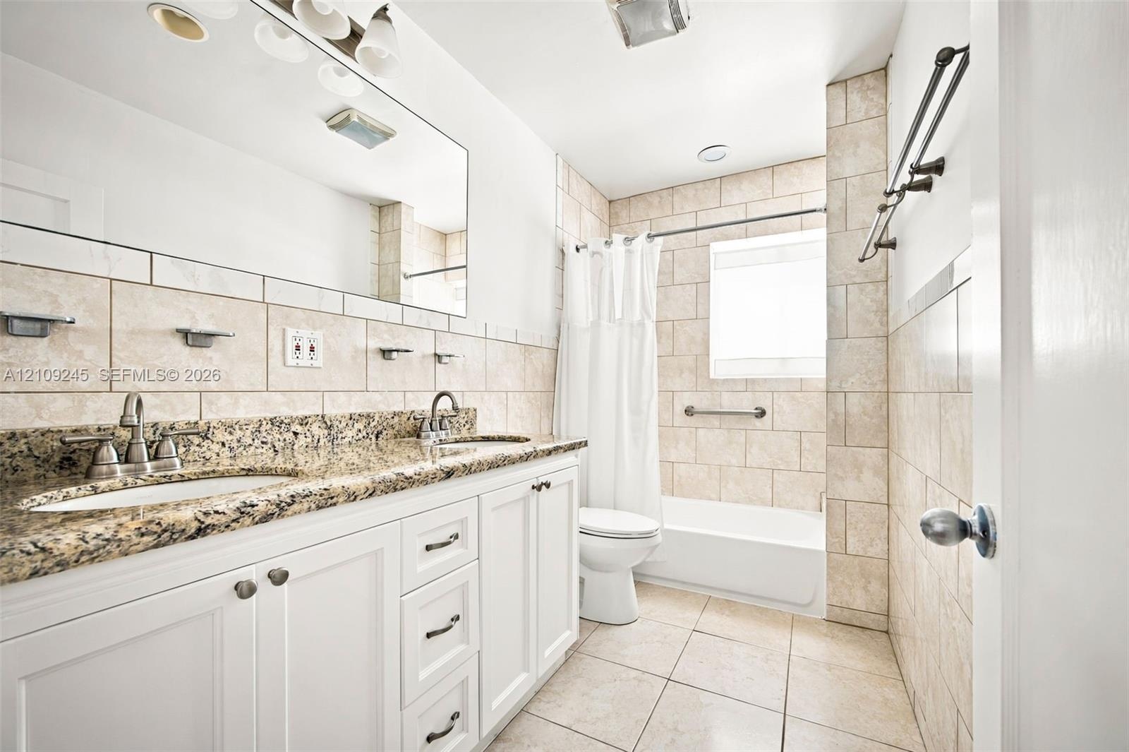 Bathroom featuring a dual-sink vanity with stone-finish countertop, white shaker-style cabinetry, tiled tub surround, and porcelain floor tiling
