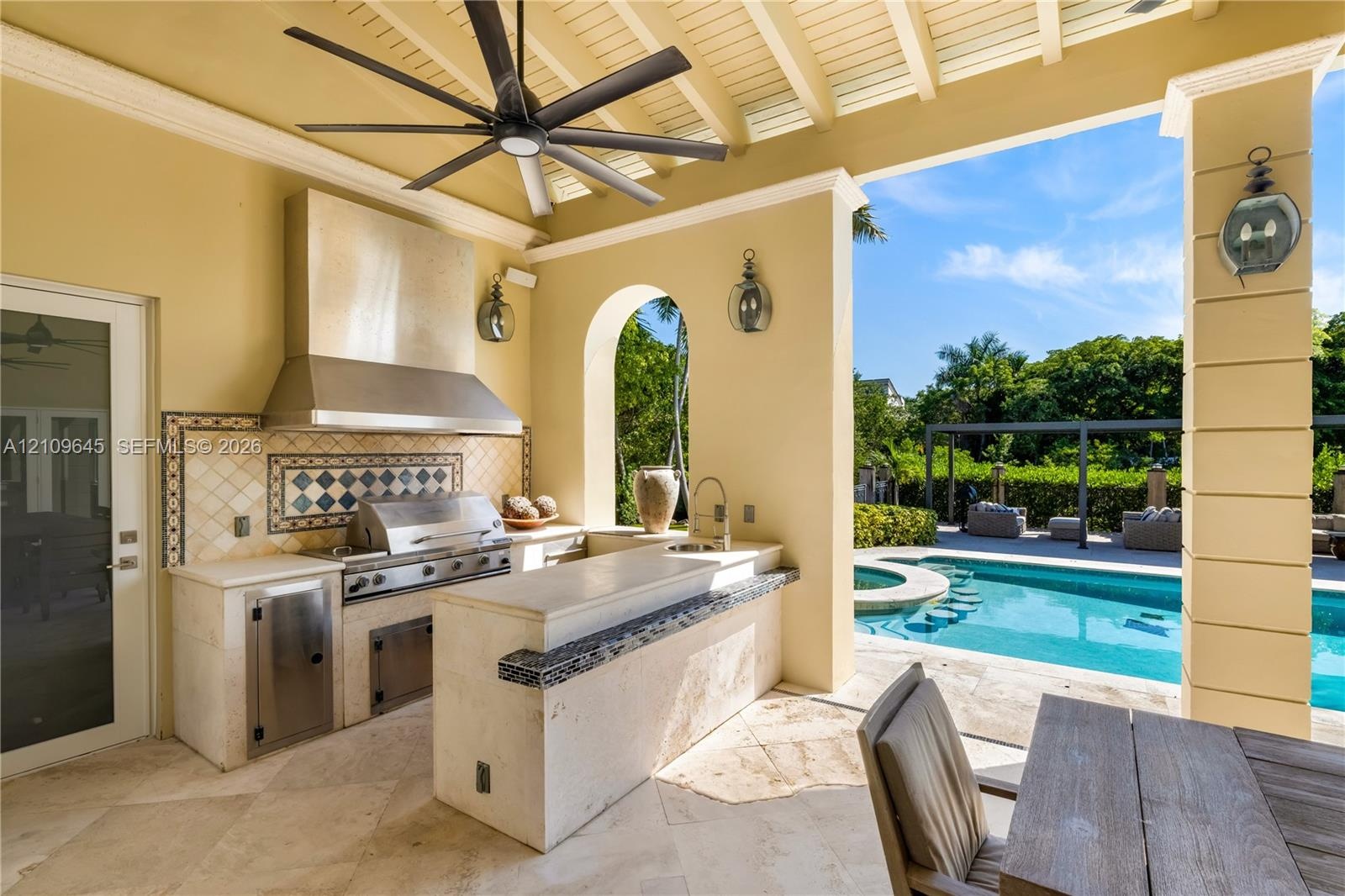 Covered outdoor kitchen featuring stone countertops, a stainless steel grill, a tile backsplash, and a built-in sink, accented by an arched opening an