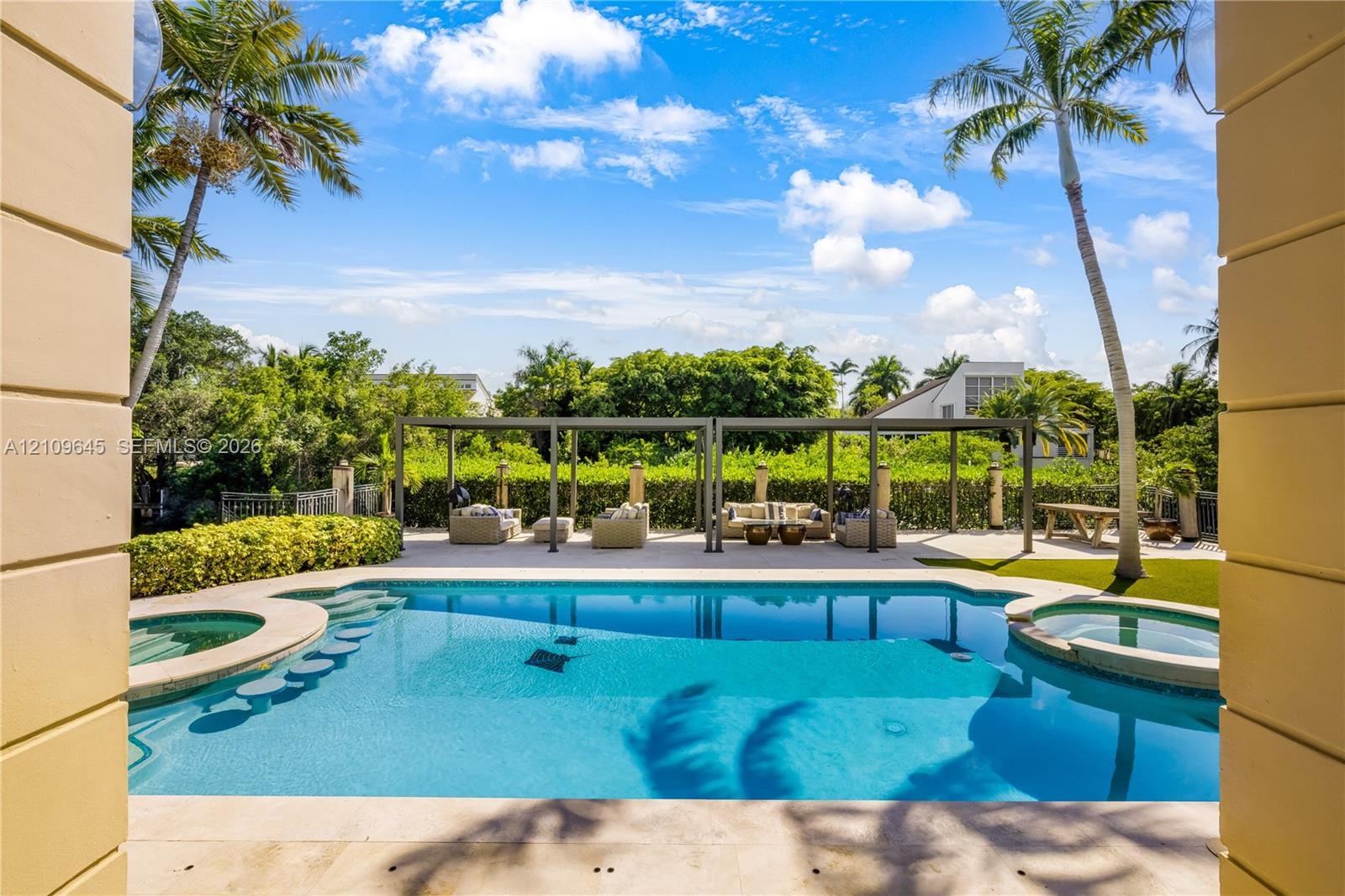 In-ground pool featuring a sun shelf, integrated spa, and surrounding stone decking, complemented by a metal pergola and mature palm trees