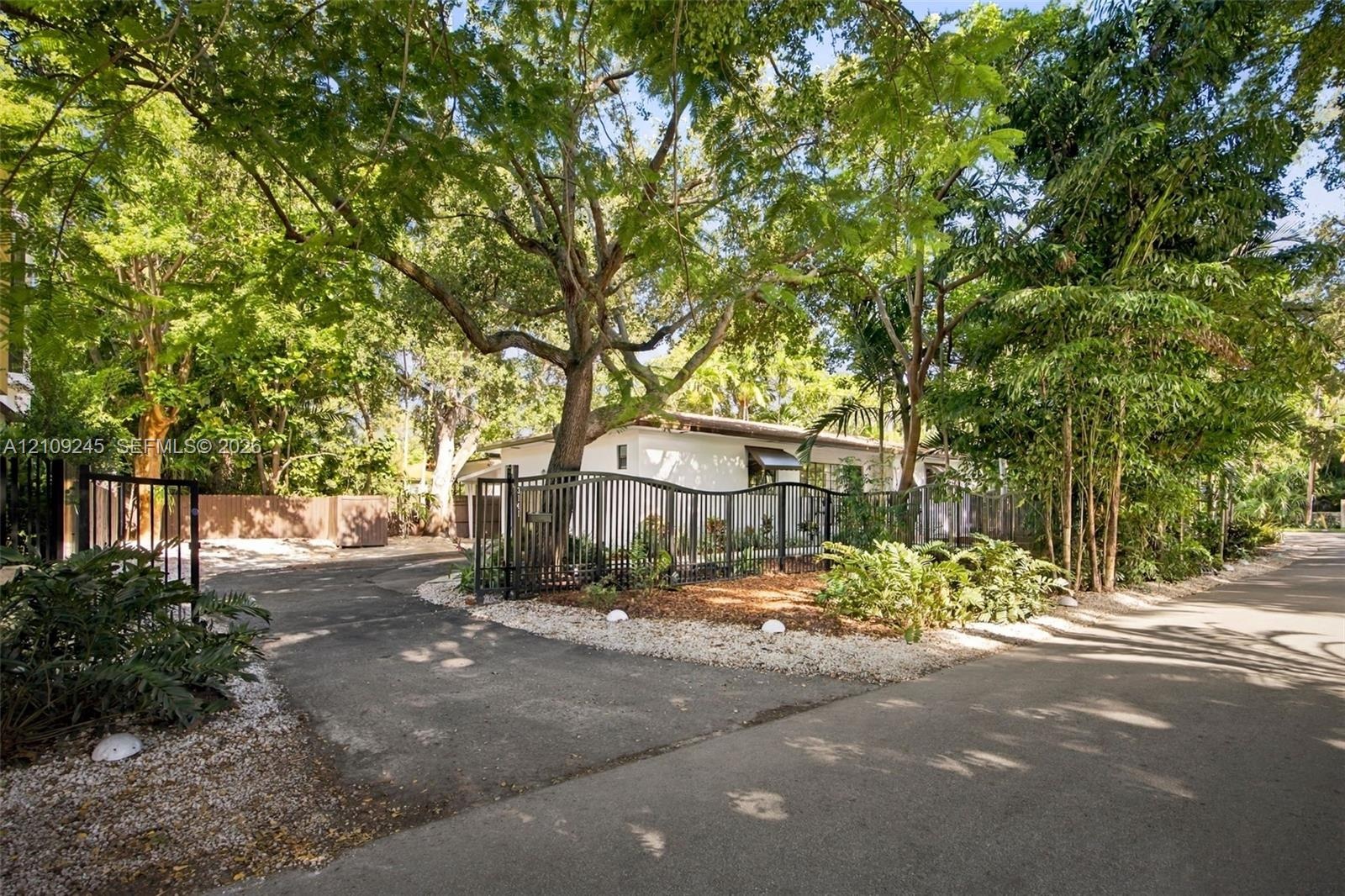 Gated entrance featuring a paved driveway, metal perimeter fencing, mature tree canopy, and landscaped stone borders