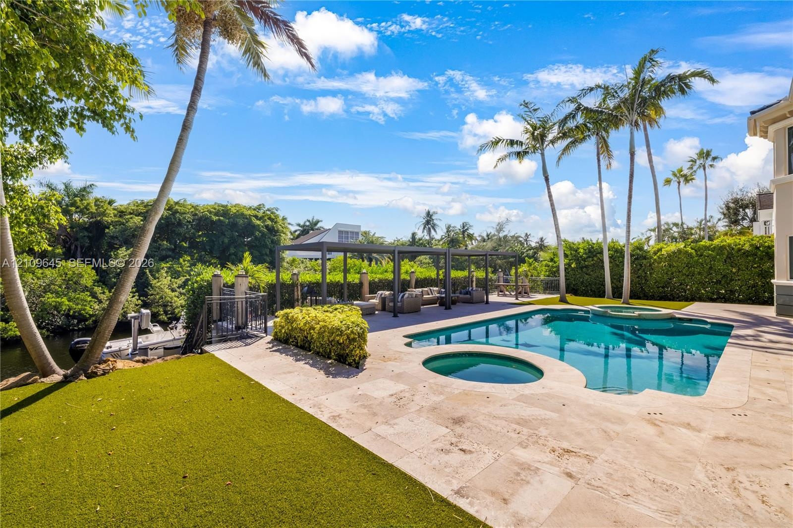 Rear yard featuring a pool with an integrated spa, stone-finish decking, a metal pergola, and synthetic turf lawn