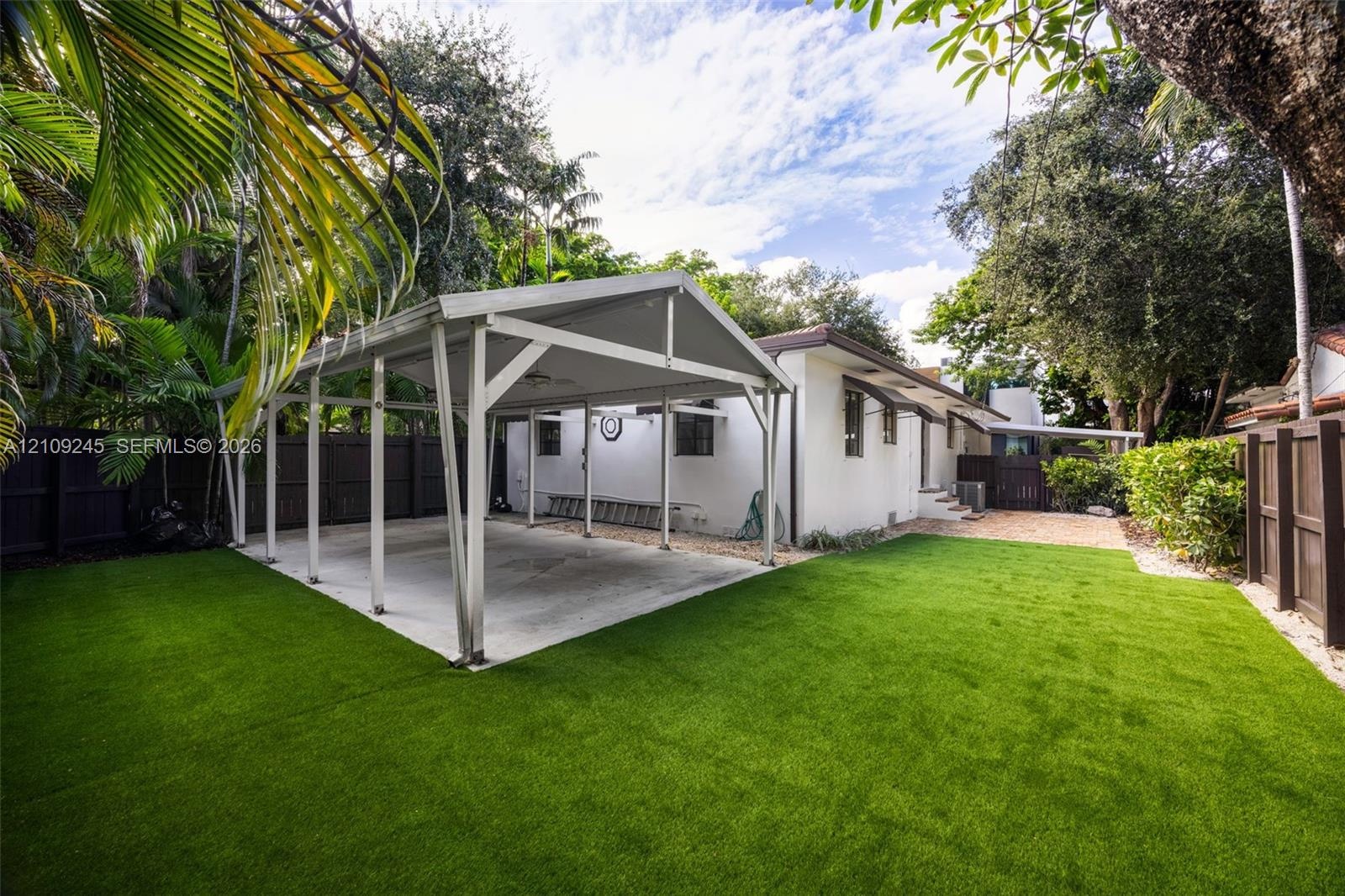 Backyard featuring a concrete-pad carport, synthetic turf lawn, dark wood-finish privacy fencing, and mature palm trees