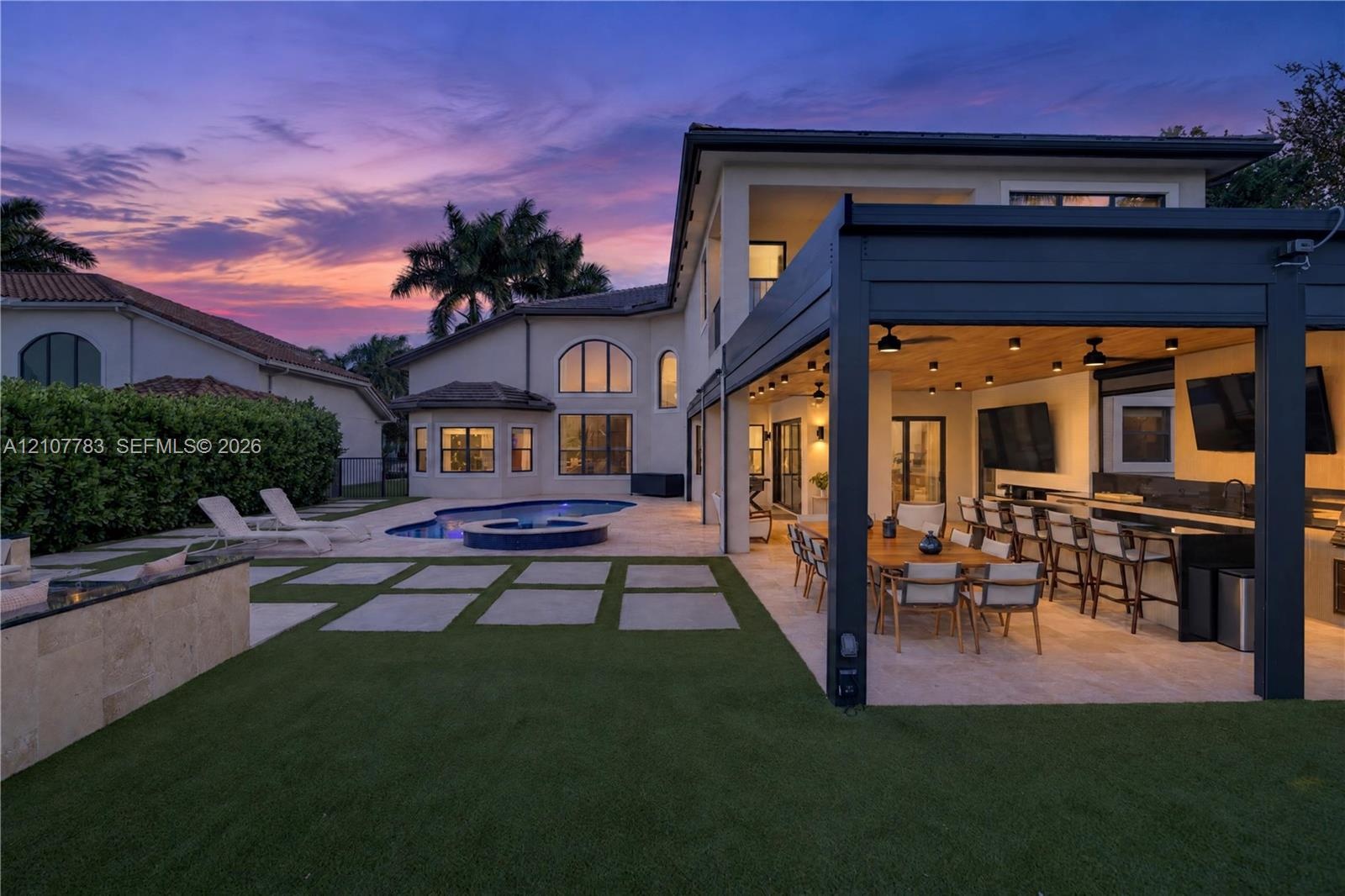 Rear exterior featuring a covered patio with a built-in outdoor kitchen, a swimming pool with an integrated spa, stone-paver hardscaping, and syntheti