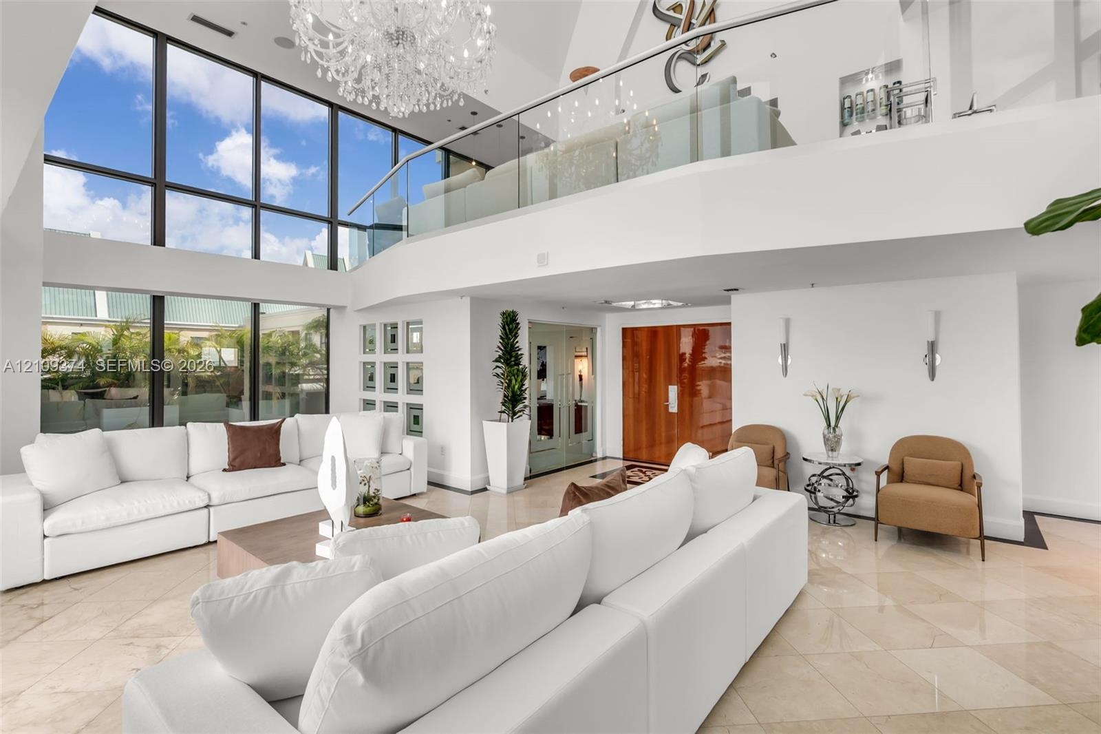 Double-height living area featuring floor-to-ceiling windows, polished stone flooring, a glass mezzanine railing, and a grand crystal chandelier