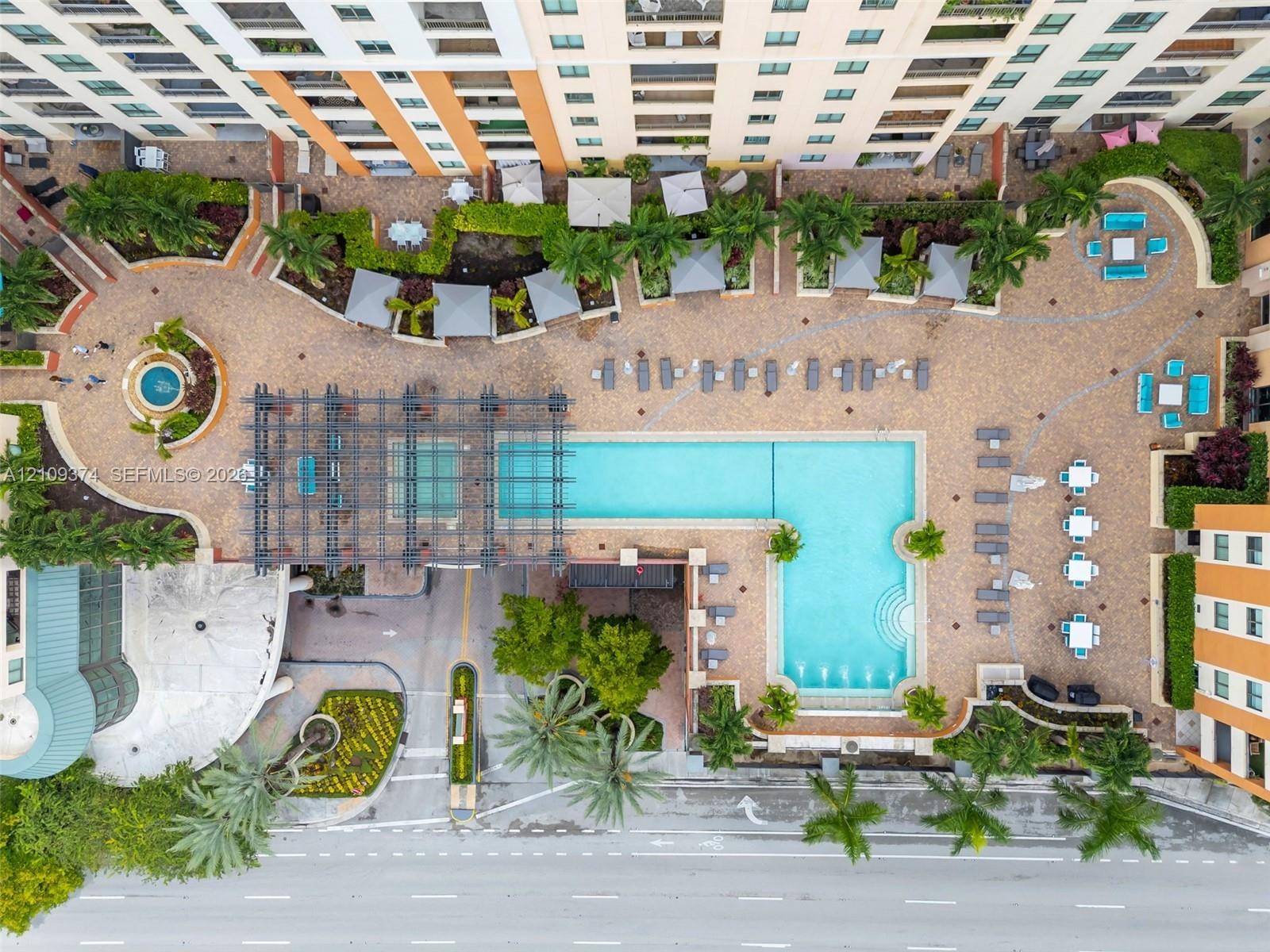 Aerial perspective of a multi-level complex featuring an L-shaped swimming pool, paved sundeck, decorative pergola, manicured landscaping, and a circu