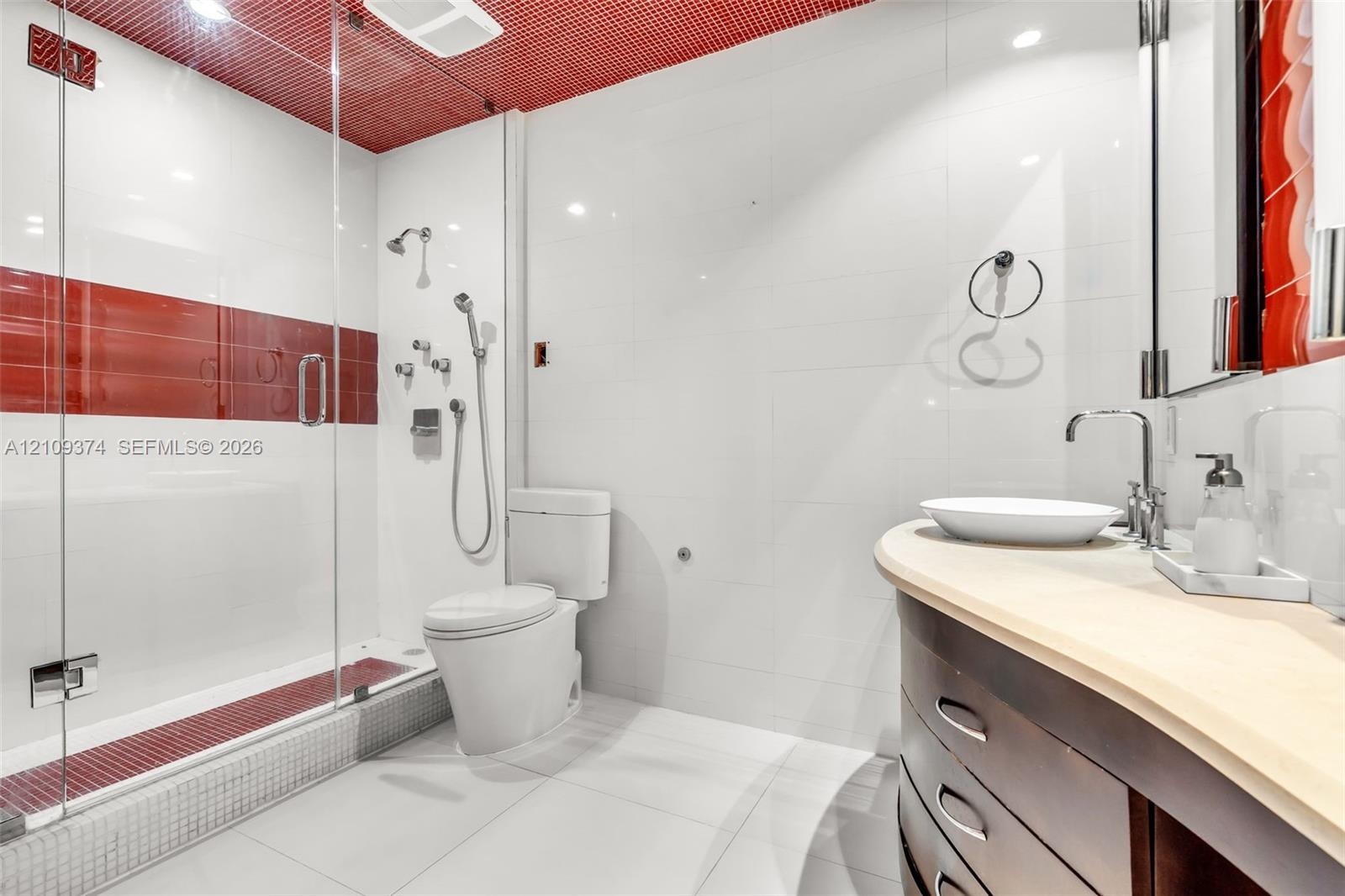 Bathroom featuring a glass-enclosed shower with multi-head fixtures and decorative tile accents, a curved wood-finish vanity with a vessel sink, and a