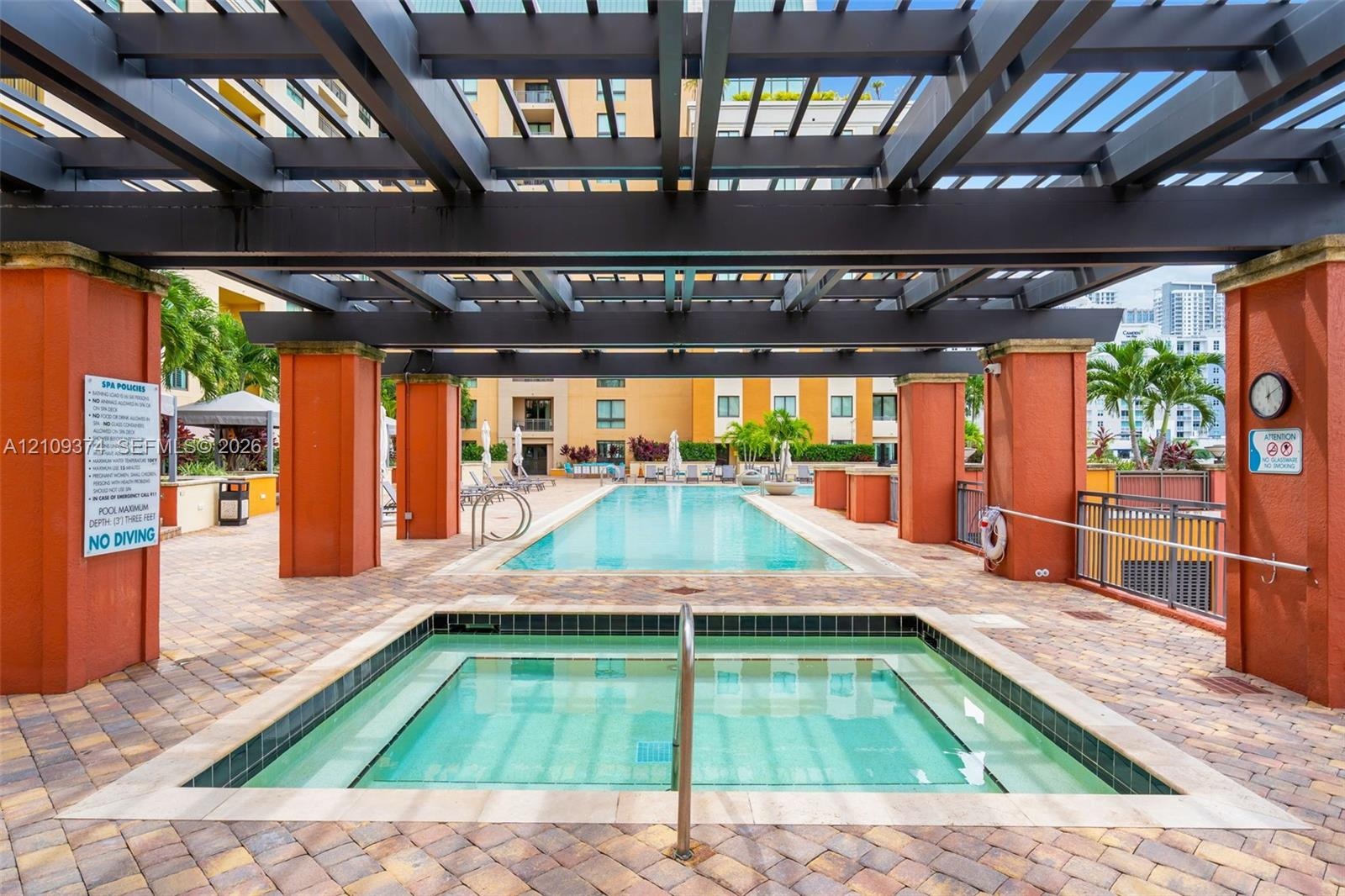 Outdoor spa and swimming pool area featuring a large overhead pergola, stone paver decking, and stucco-finished columns