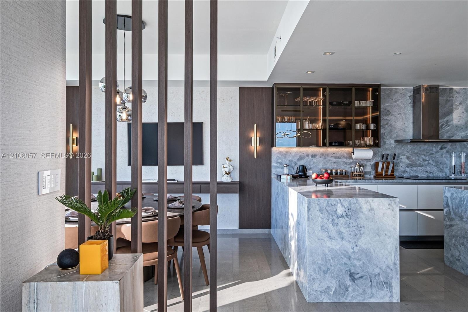 Kitchen featuring stone-clad waterfall island and backsplash, custom cabinetry with glass-front uppers, decorative vertical wood slat room divider, an