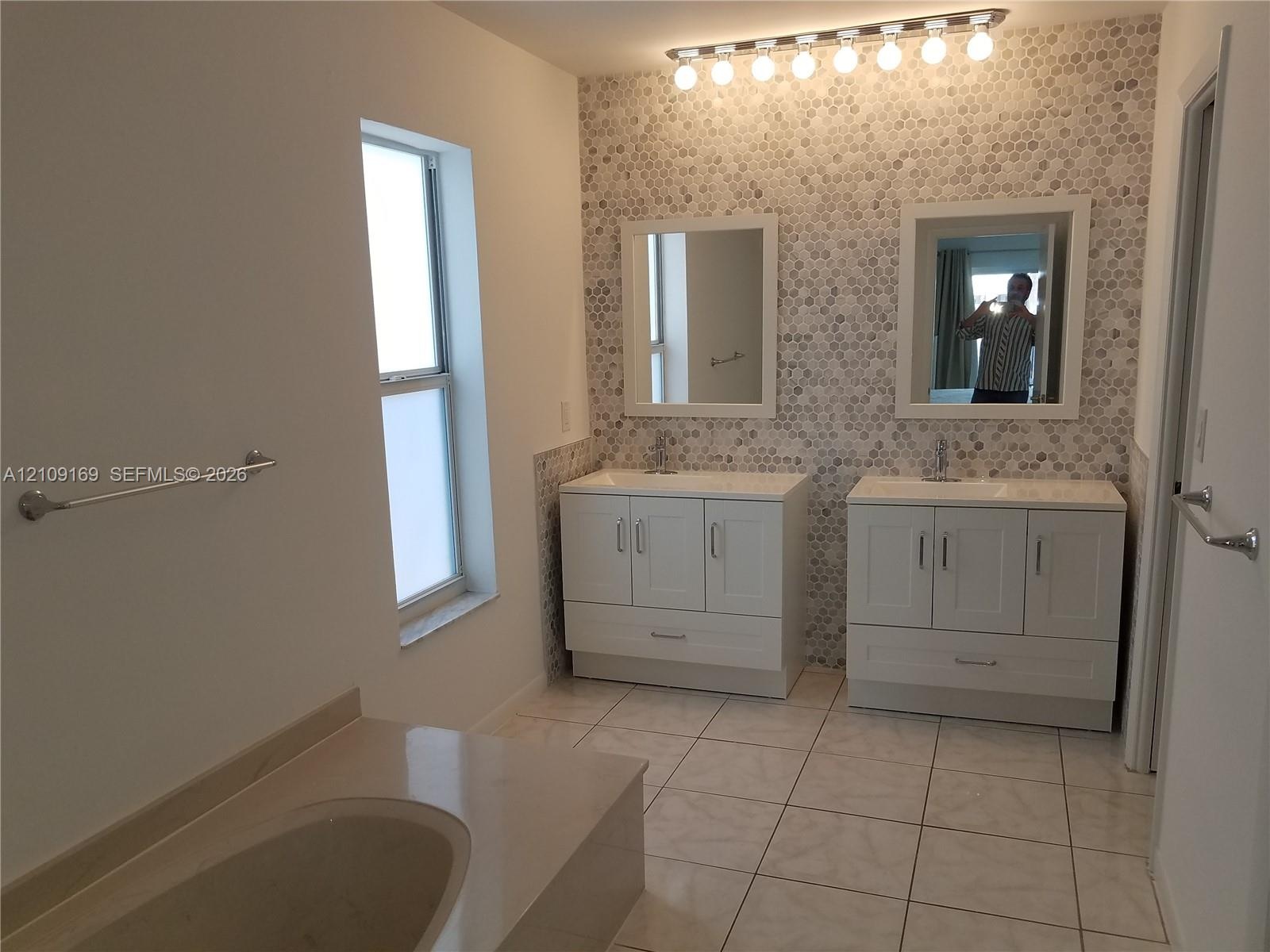 Dual white vanities with integrated sinks, penny-round tile backsplash, tiled flooring, built-in soaking tub, and matching rectangular wall mirrors