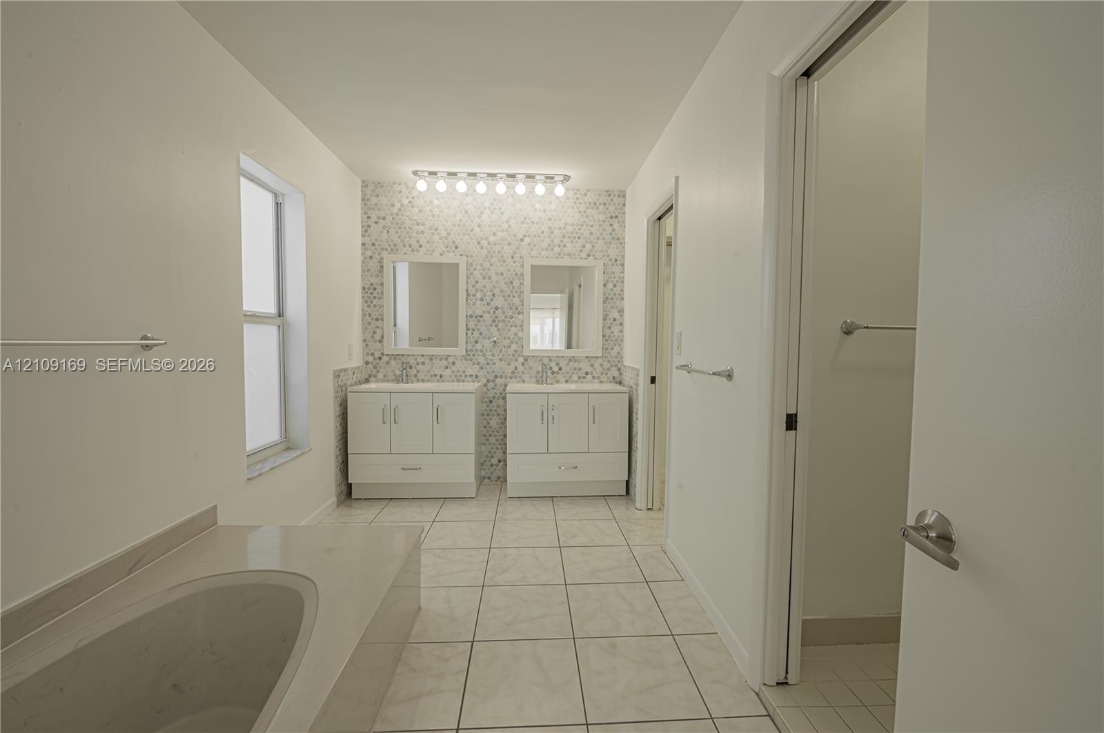 Dual white vanities, multi-colored stone-tiled accent wall, built-in soaking tub, large-format tile flooring, and multi-bulb integrated vanity lightin