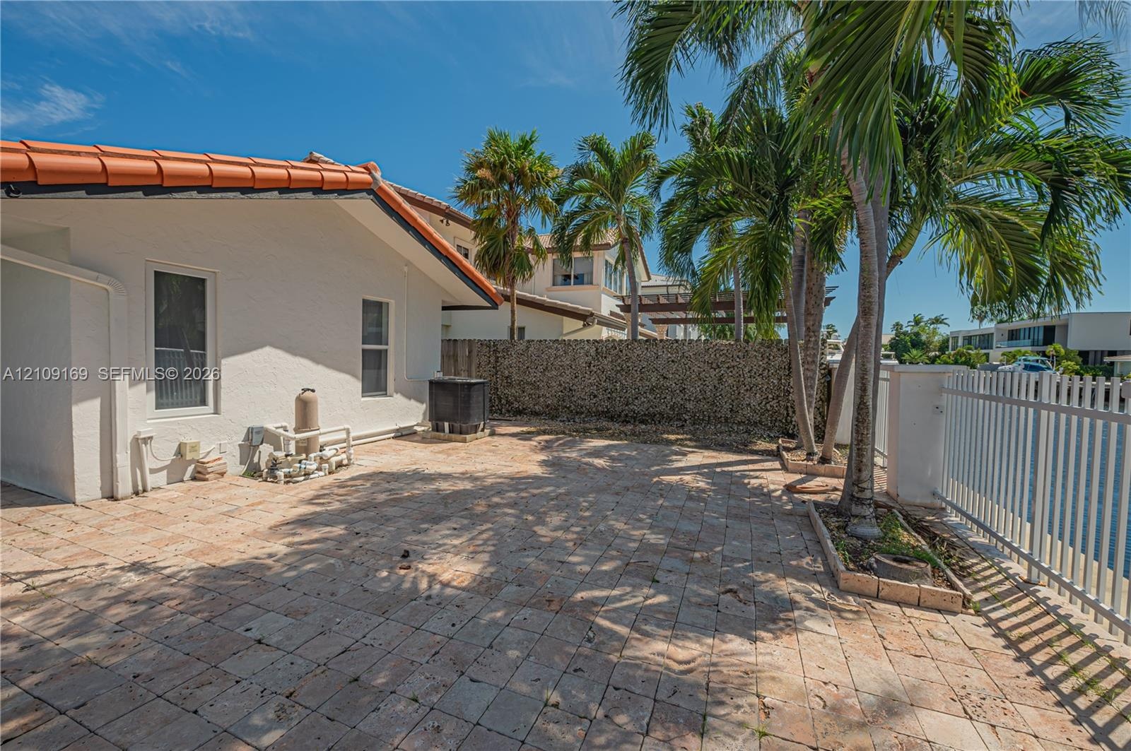 Rear exterior patio featuring a light-toned paver surface, white metal fencing, and mature palm trees