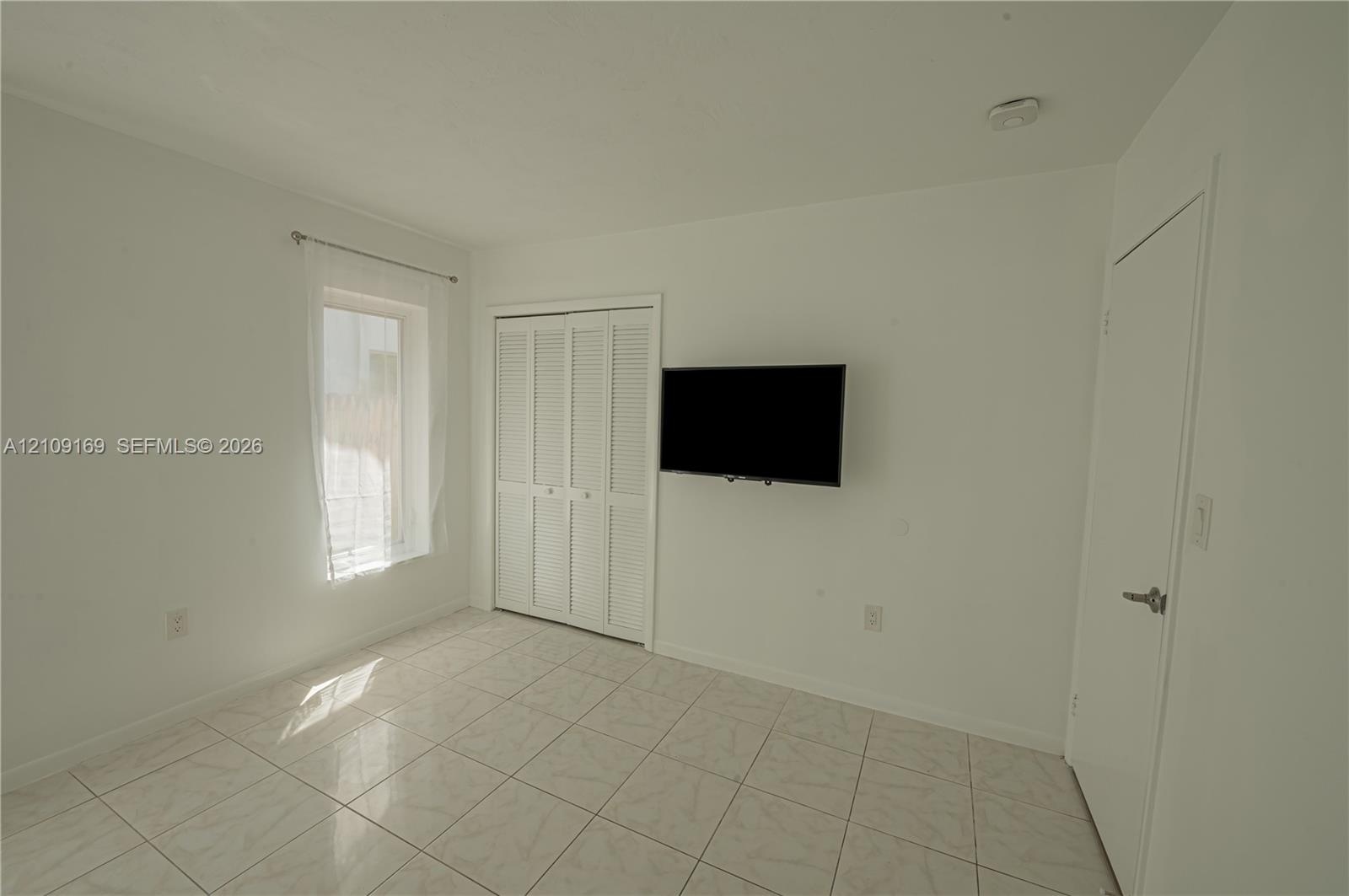 Interior space featuring large-format ceramic tile flooring, a window with a metal curtain rod, and a built-in closet with white louvered bi-fold door