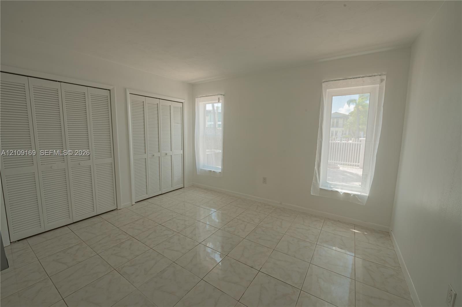 Interior space featuring neutral tile flooring, two sets of white louvered-door closets, and dual windows
