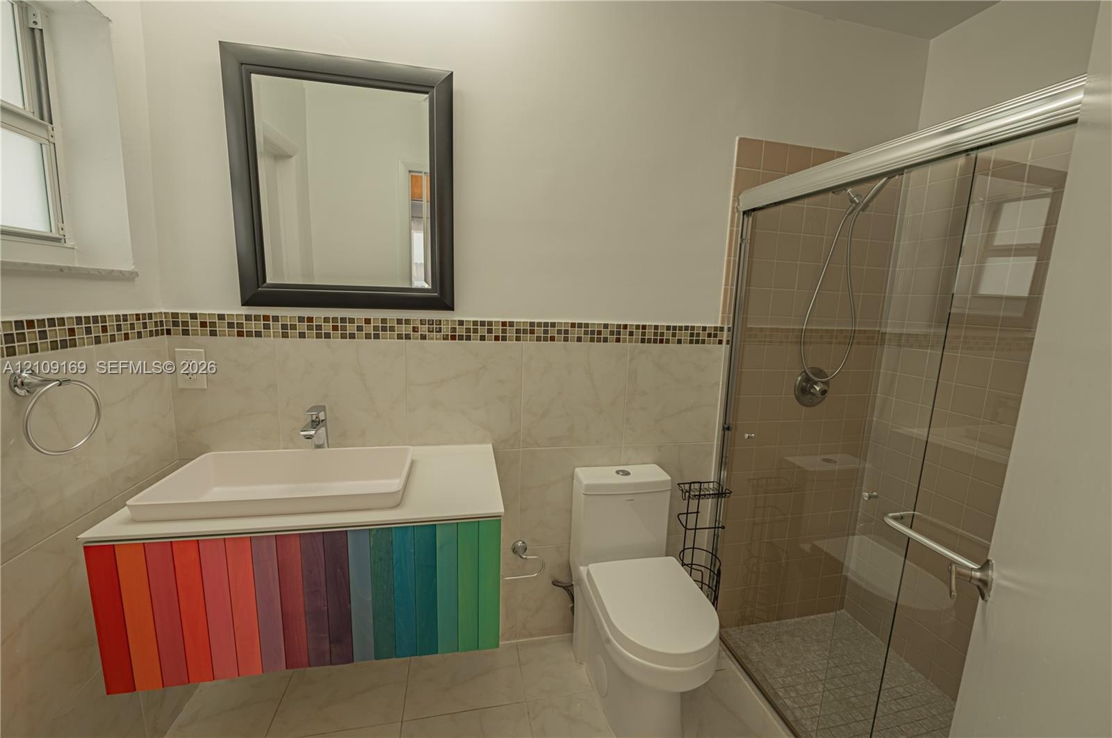 Bathroom featuring a floating vanity with a multicolored cabinet base, rectangular vessel sink, mosaic tile accent border, and a glass-enclosed shower